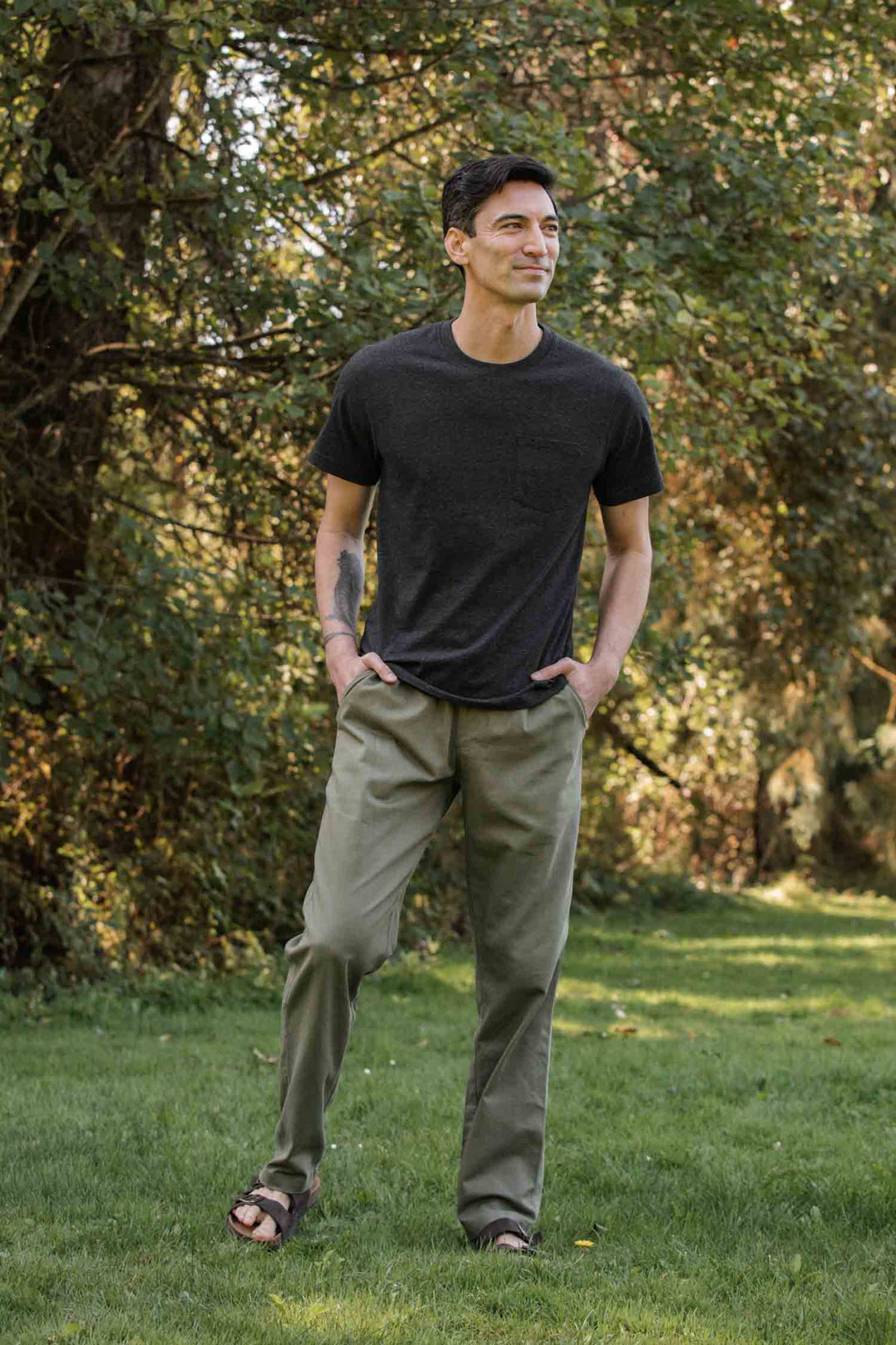 A man stands outdoors on grass, wearing the Bridge & Burn Recycled Pocket Tee in Charcoal Heather, paired with olive green pants and sandals, hands in his pockets, with trees visible in the background.