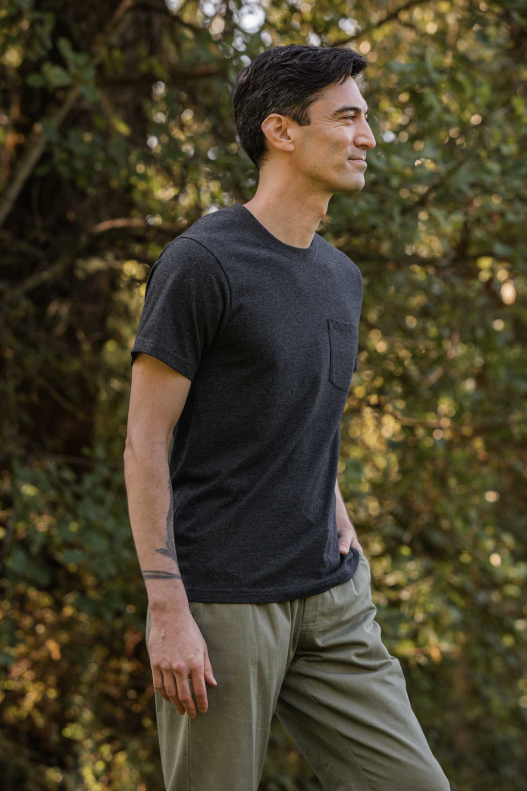 A man with short dark hair wears a Bridge & Burn Recycled Pocket Tee in Charcoal Heather and light green pants, standing outdoors with one hand in his pocket, surrounded by greenery and sunlight.