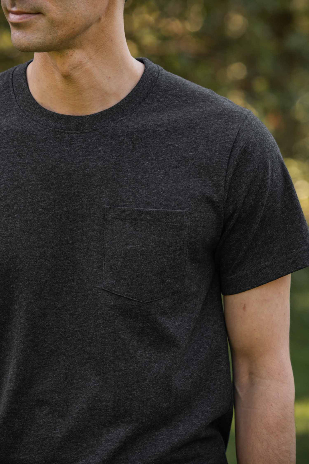 Someone wearing the Bridge & Burn Recycled Pocket Tee in Charcoal Heather, featuring a chest pocket and made from a recycled cotton blend, stands outdoors among blurred greenery with only their lower face and upper torso visible.