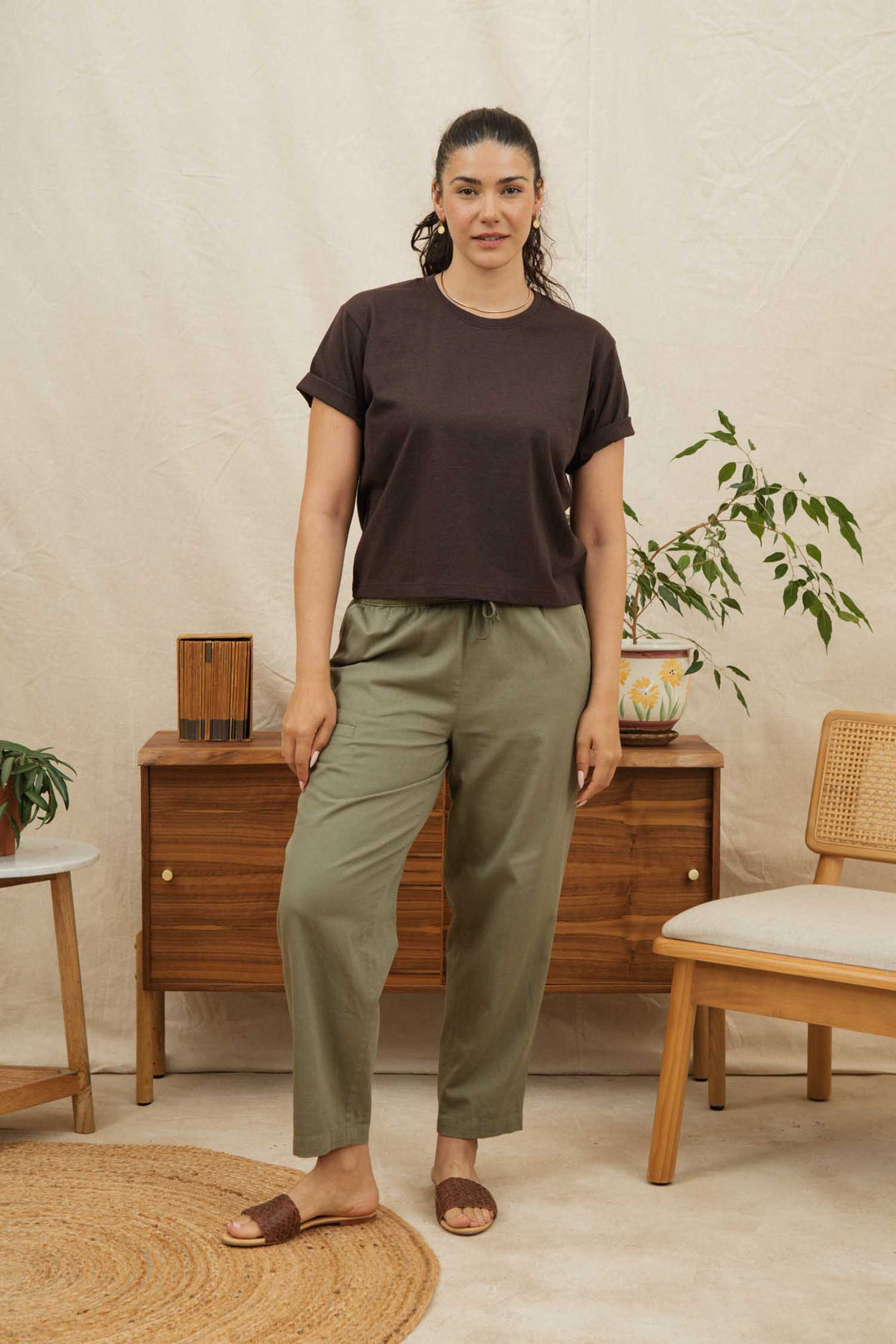 A woman stands indoors wearing the Bridge & Burn Recycled Boxy Tee in Truffle, olive green pants, and brown sandals. She is by a wooden dresser with plants and decor, beside a chair and round rug on a light floor.
