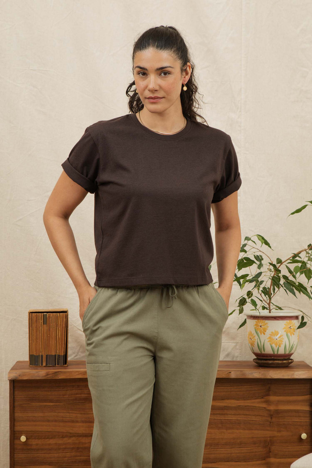 A woman with dark hair in a ponytail wears the Bridge & Burn Recycled Boxy Tee in Truffle and olive green pants, standing hands-in-pockets before a wooden cabinet with a potted plant and small decorative box.