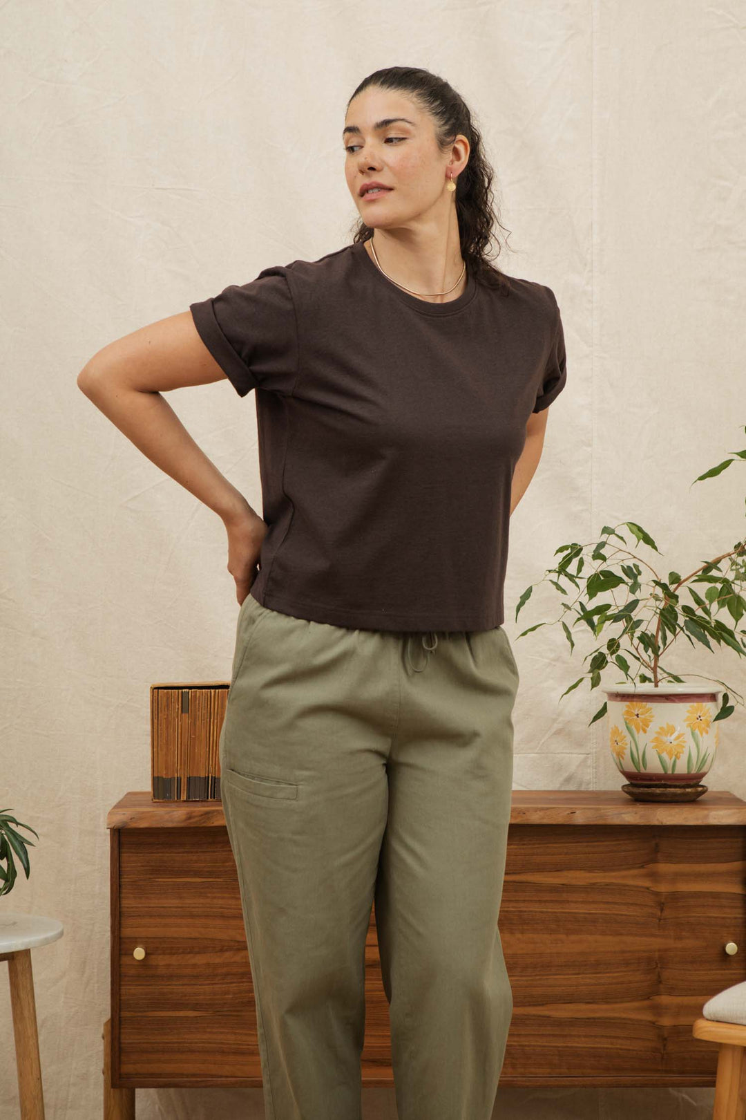 A woman stands indoors, hands behind her back, wearing the Bridge & Burn Recycled Boxy Tee in Truffle and olive green pants. She is in front of a wooden cabinet with a potted plant and a decorative box against a light fabric backdrop.
