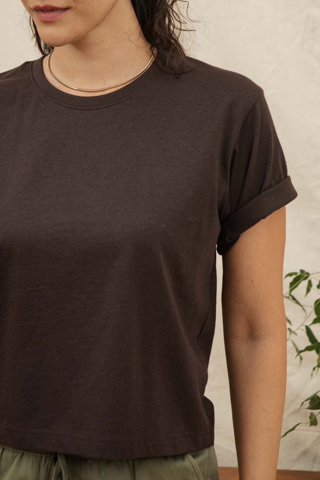 A person wears the Bridge & Burn Recycled Boxy Tee in Truffle—a dark brown vintage-style top with rolled cuffs—paired with a simple necklace, indoors by a plant. Only their upper torso and part of their face are visible.
