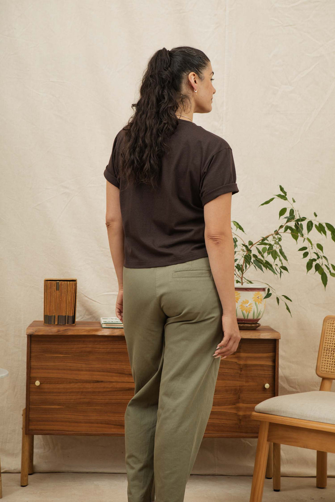 A woman with long dark hair in a ponytail stands facing away from the camera, wearing the Bridge & Burn Recycled Boxy Tee in Truffle and olive green pants, in a room with wooden furniture and a potted plant.