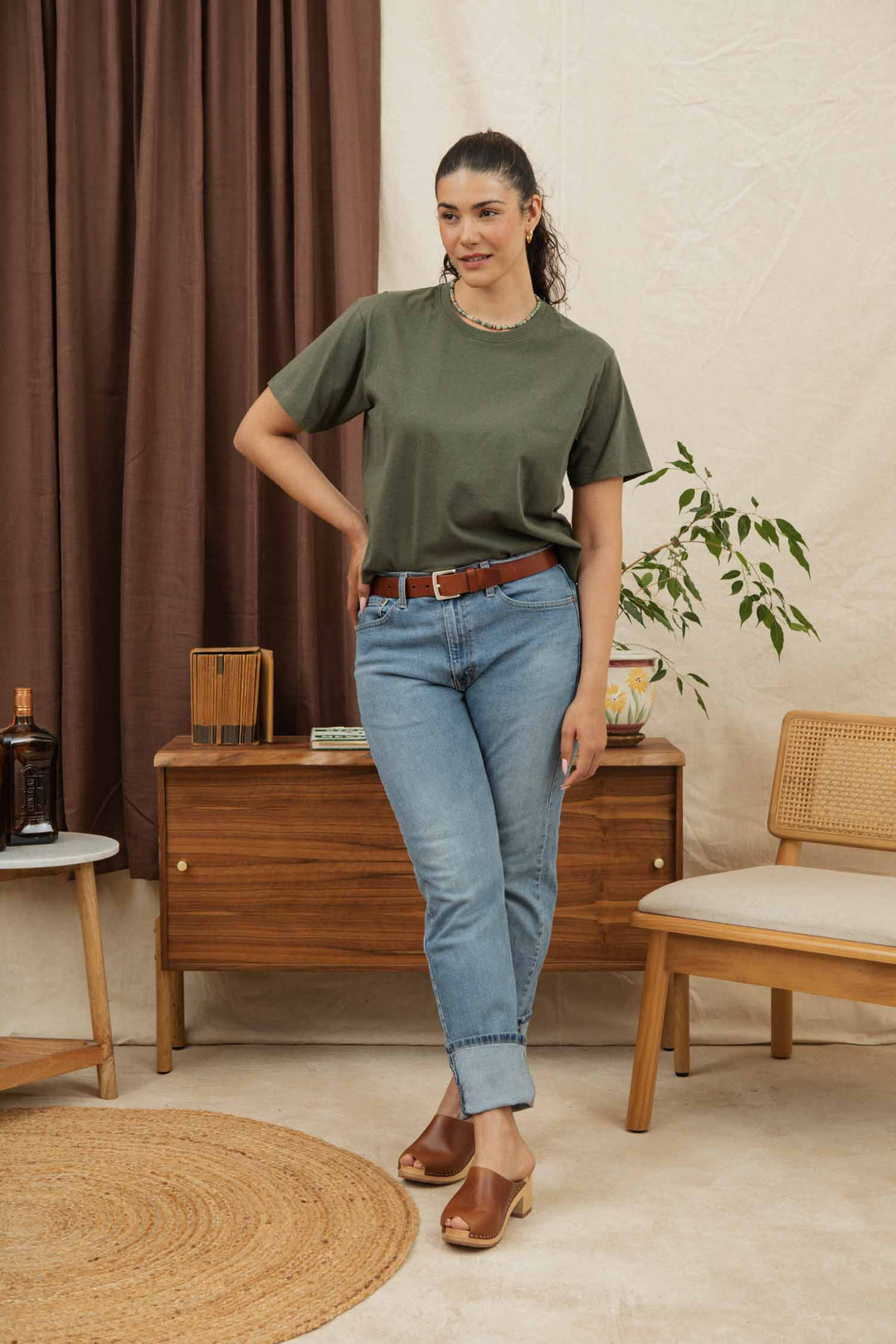 A woman indoors wears the Bridge & Burn Recycled Boxy Tee in Olive with blue jeans, a brown belt, and clogs. She poses by a wooden sideboard, a plant, and a chair, with brown curtains in the background.