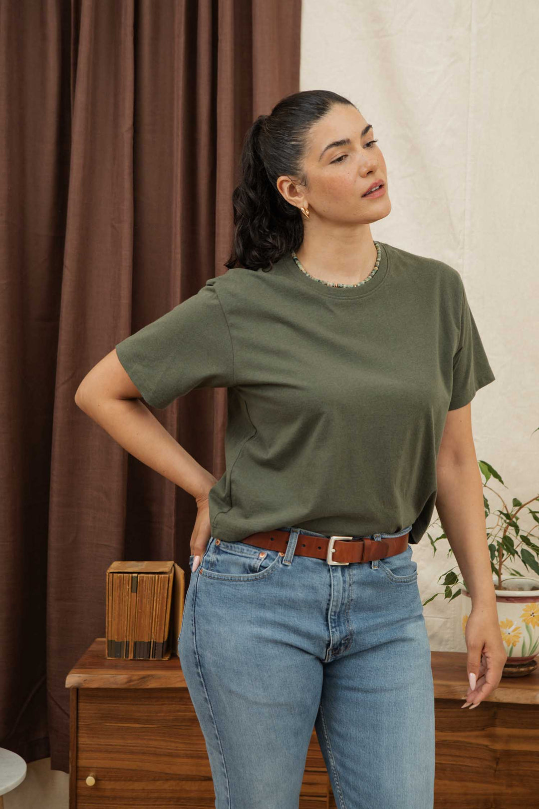 A woman with dark hair in a ponytail stands indoors wearing the Bridge & Burn Recycled Boxy Tee in olive, blue jeans, and a brown belt. She rests her hand on her lower back beside a wooden cabinet with a potted plant and brown curtain.