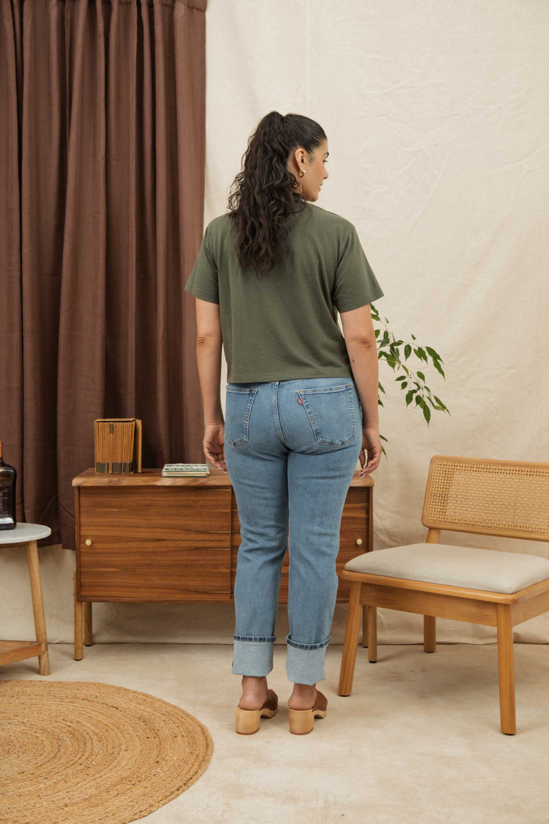 A woman with long dark hair in a ponytail stands facing away, wearing the Bridge & Burn Recycled Boxy Tee in Olive, blue jeans, and light heeled clogs in a mid-century modern room with wood furniture and neutral decor.