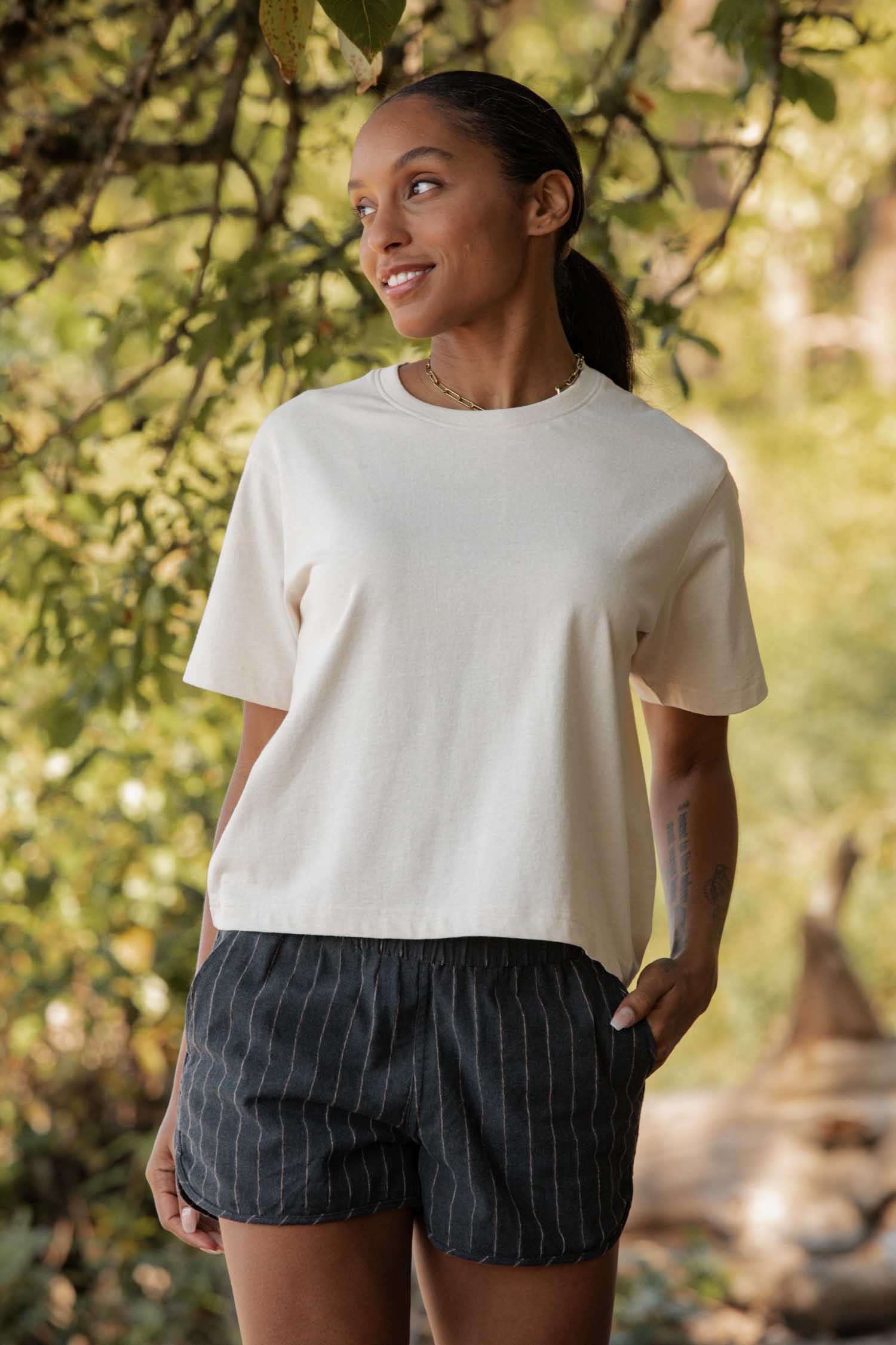 A woman stands outdoors under leafy branches, smiling and looking to the side. She wears the Bridge & Burn Recycled Boxy Tee in Ivory with dark pinstriped shorts, one hand in her pocket as sunlight filters through the trees behind her.