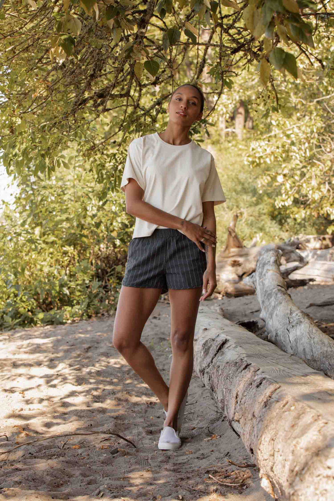 A woman stands on a wooded path beside a fallen tree, wearing the Bridge & Burn Recycled Boxy Tee in Ivory, striped shorts, and white sneakers as sunlight filters through green leaves above her.