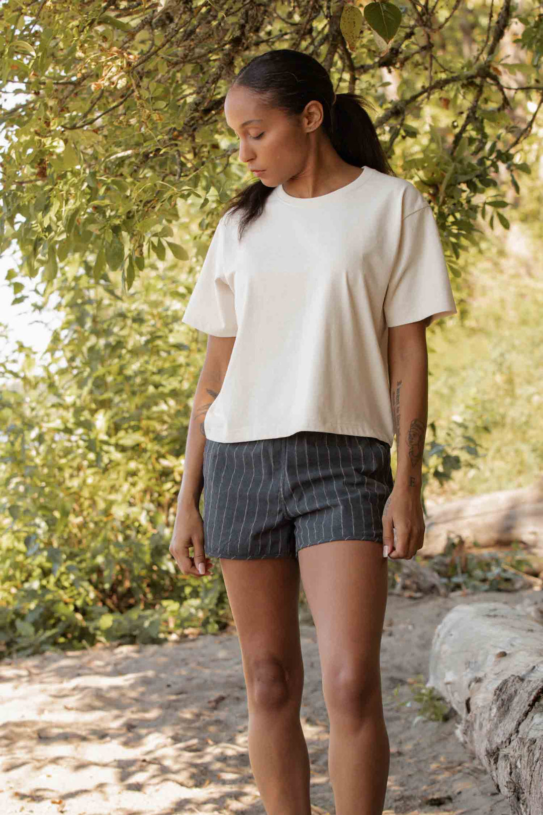 Under sun-dappled trees, someone stands wearing the Bridge & Burn Recycled Boxy Tee in Ivory and dark pinstriped shorts, hands by their sides as sunlight creates a natural glow through the leaves.
