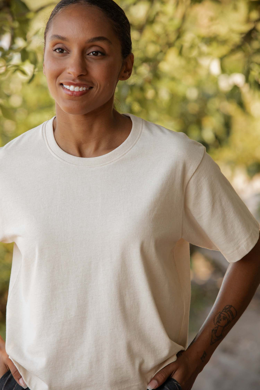 A person wearing the Bridge & Burn Recycled Boxy Tee in Ivory stands outdoors, smiling with hands on hips. Green foliage and sunlight blur in the background, and a small tattoo is visible on their left forearm.