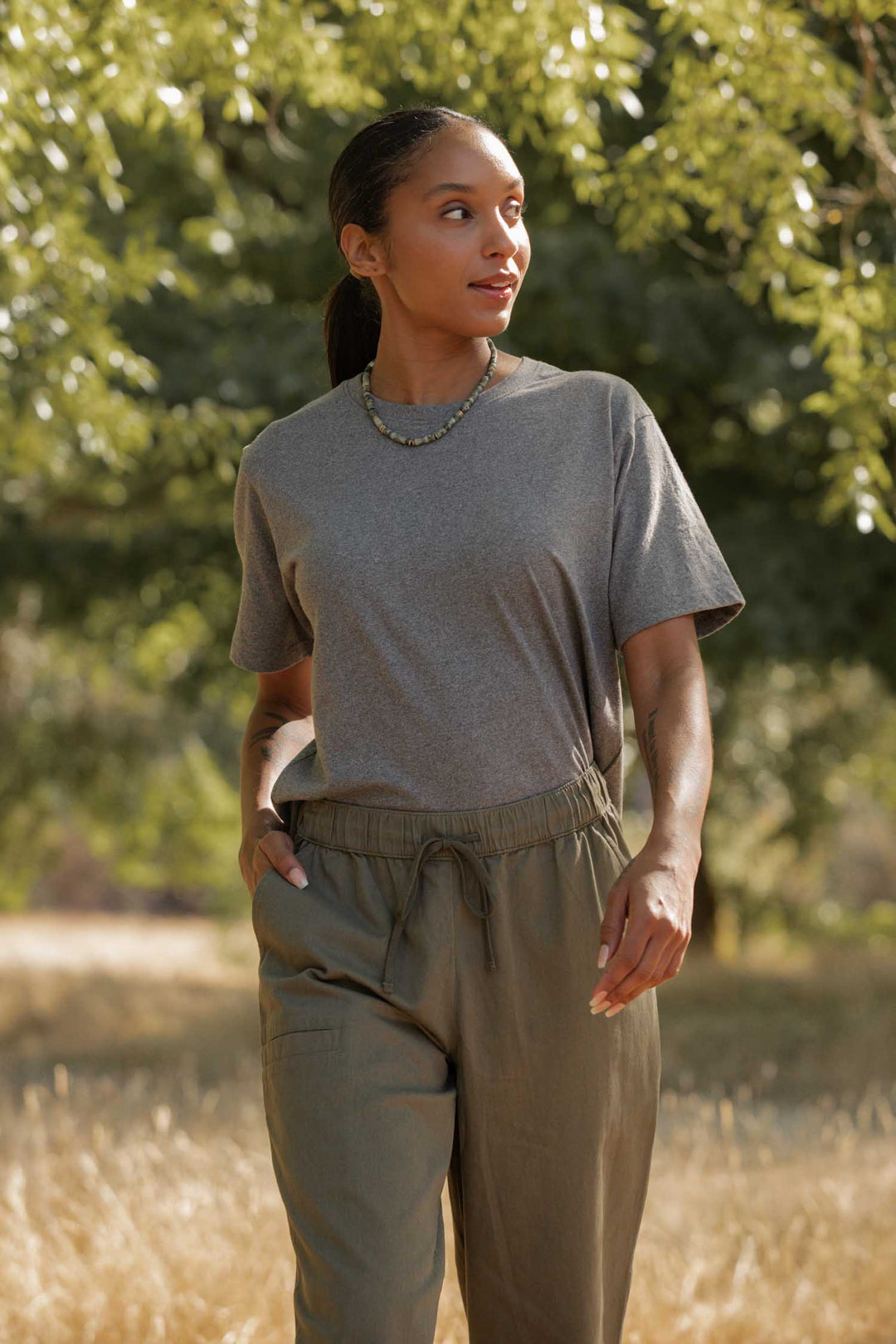 A woman wears the Bridge & Burn Recycled Boxy Tee in Grey Heather with olive green pants as she walks outdoors in a grassy, sunlit area with trees, gazing to the side with a neutral expression.