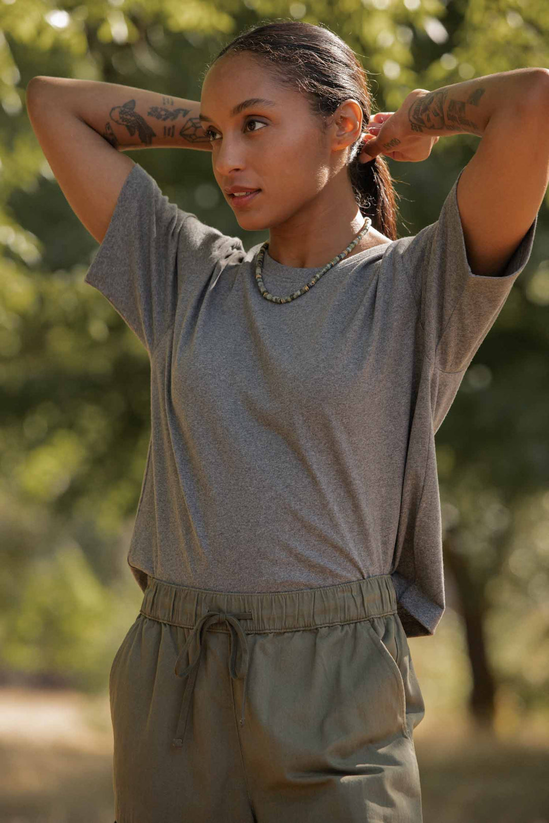 A tattooed woman ties her hair back outdoors, wearing the Bridge & Burn Recycled Boxy Tee in Grey Heather, olive green pants, and a beaded necklace amid sunlit trees.
