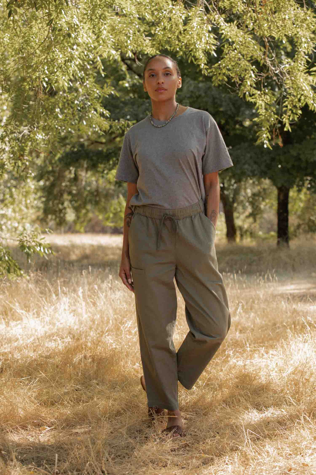 A person wears the Bridge & Burn Recycled Boxy Tee in Grey Heather and olive pants, standing on dry grass under leafy trees. Sunlight filters through the branches as they face the camera with a neutral expression.