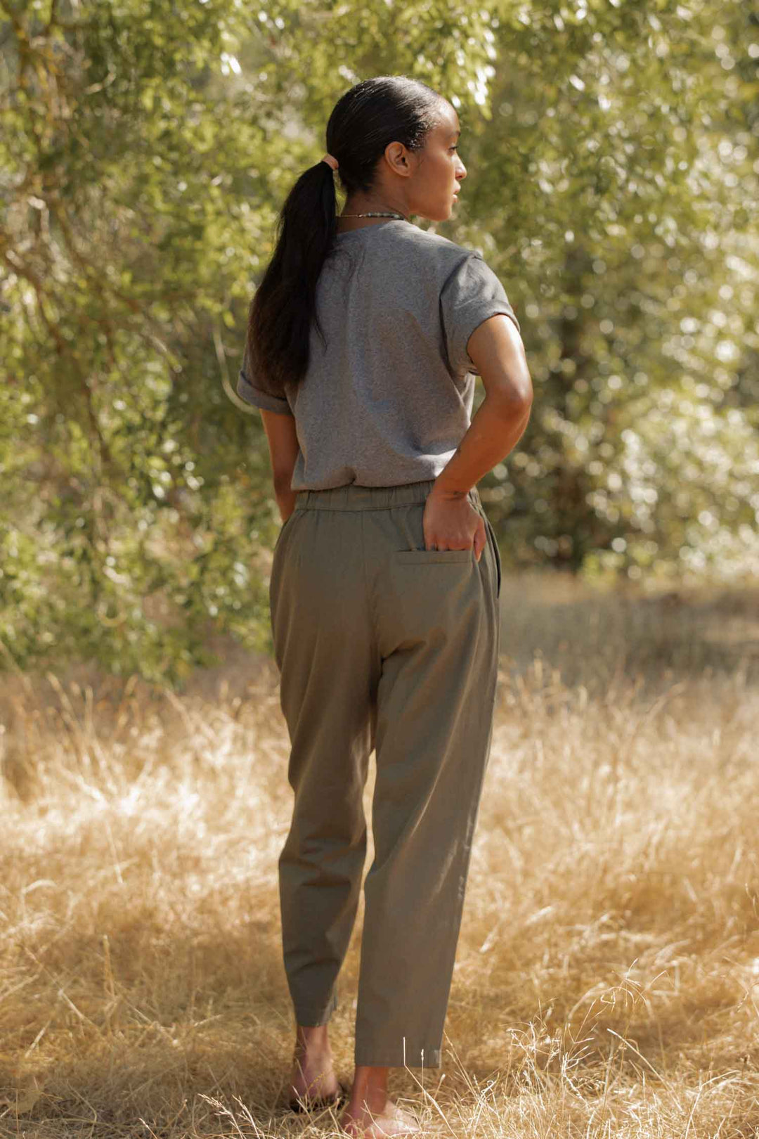 A woman with a ponytail stands outside on dry grass, facing away, wearing the Bridge & Burn Recycled Boxy Tee in Grey Heather and olive-green pants, one hand in her back pocket with trees in the background.