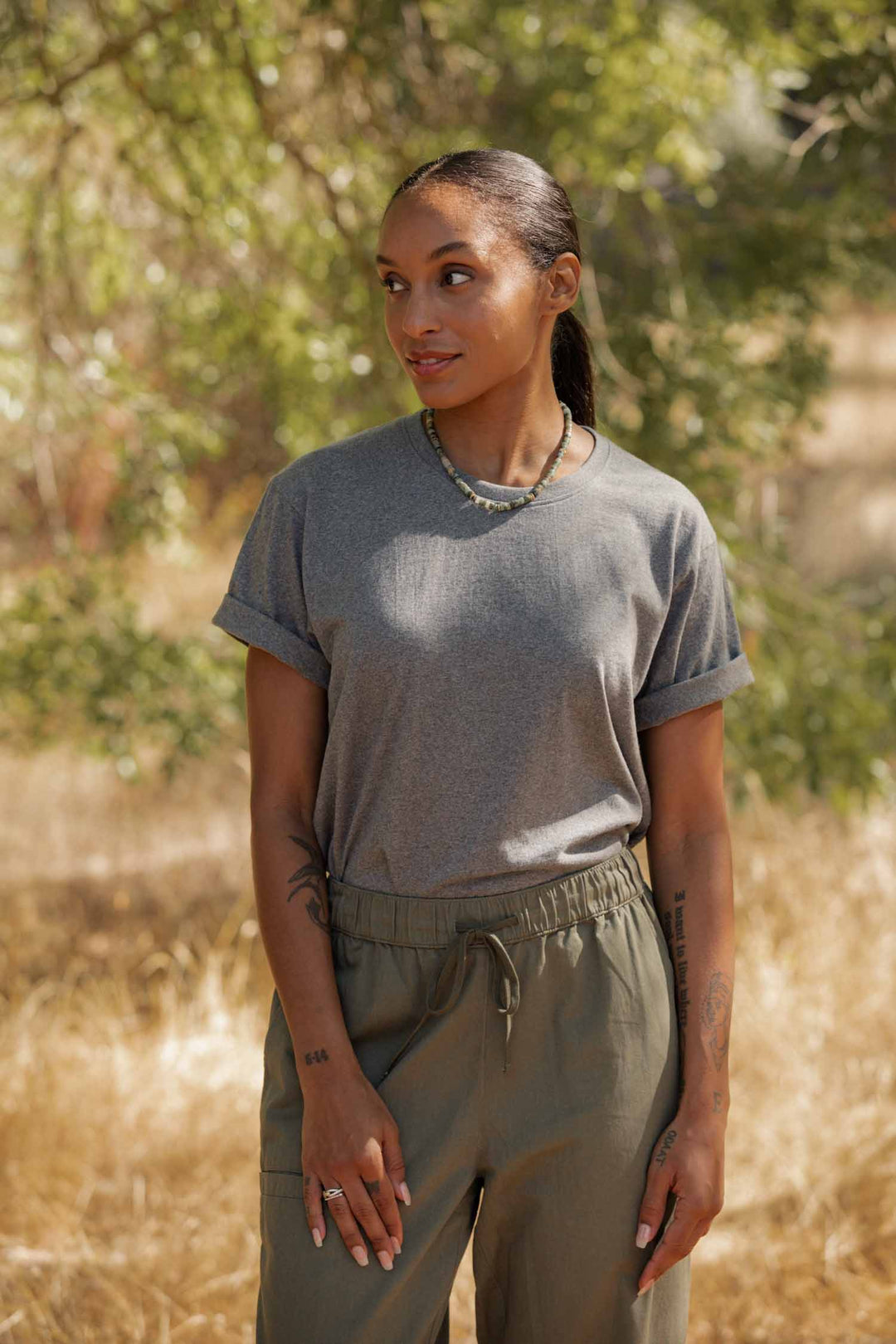 A woman with long braided hair and arm tattoos stands outdoors among trees, wearing the Bridge & Burn Recycled Boxy Tee in Grey Heather and olive green pants. Sunlight highlights her sustainable style as she looks slightly to the side.