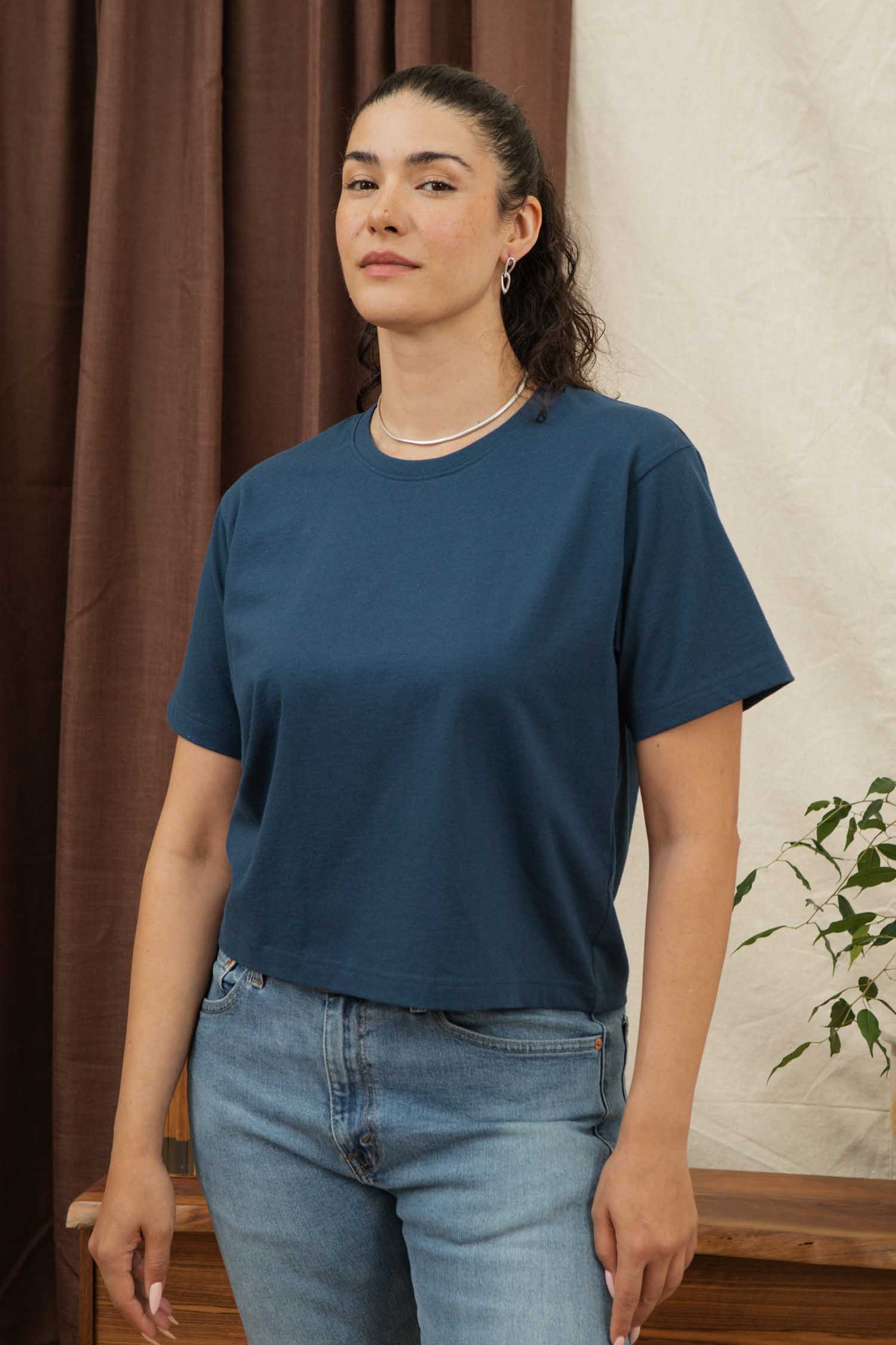 A woman with dark, wavy hair pulled back stands indoors by brown and cream curtains, wearing the Bridge & Burn Recycled Boxy Tee in Cobalt with light blue jeans. A small plant is visible on her right.