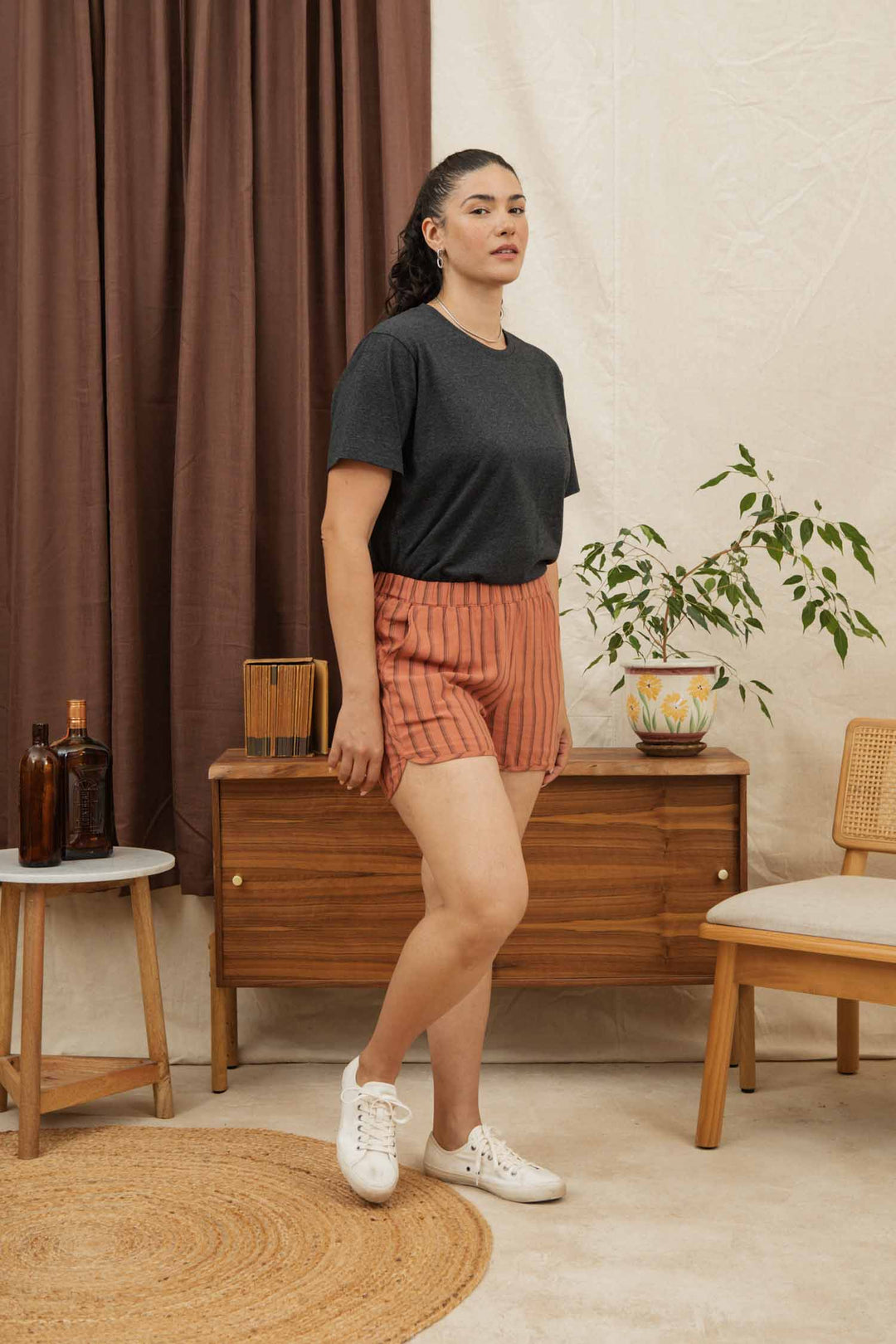A woman stands indoors wearing the Bridge & Burn Recycled Boxy Tee in Charcoal Heather, paired with orange striped shorts and white sneakers. She is near a wooden sideboard with decor, a potted plant, and brown curtains behind her.