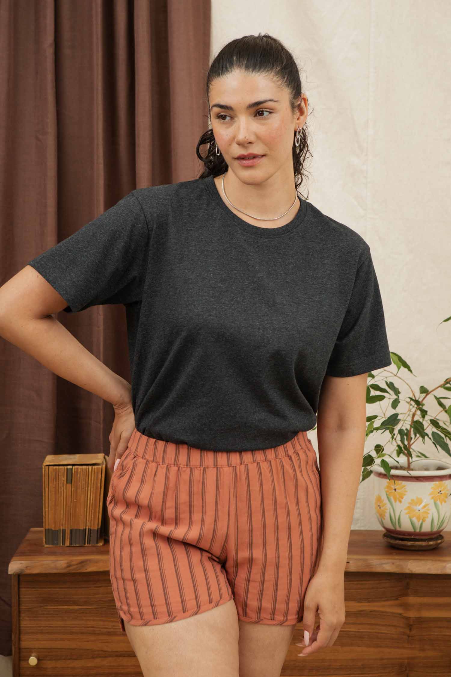 A woman with dark hair in a ponytail stands indoors, confidently wearing the Bridge & Burn Recycled Boxy Tee in Charcoal Heather, paired with orange striped shorts near a wooden table topped with a potted plant and books.