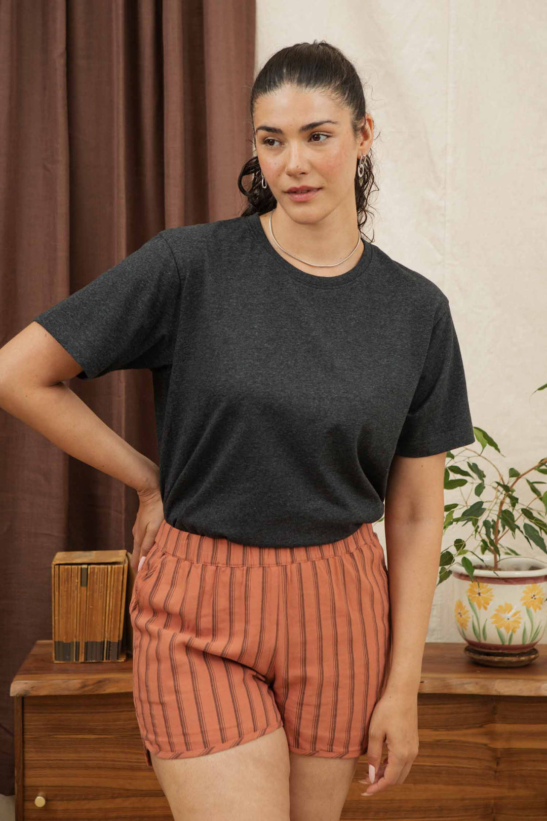 A woman with dark hair in a ponytail stands indoors, confidently wearing the Bridge & Burn Recycled Boxy Tee in Charcoal Heather, paired with orange striped shorts near a wooden table topped with a potted plant and books.