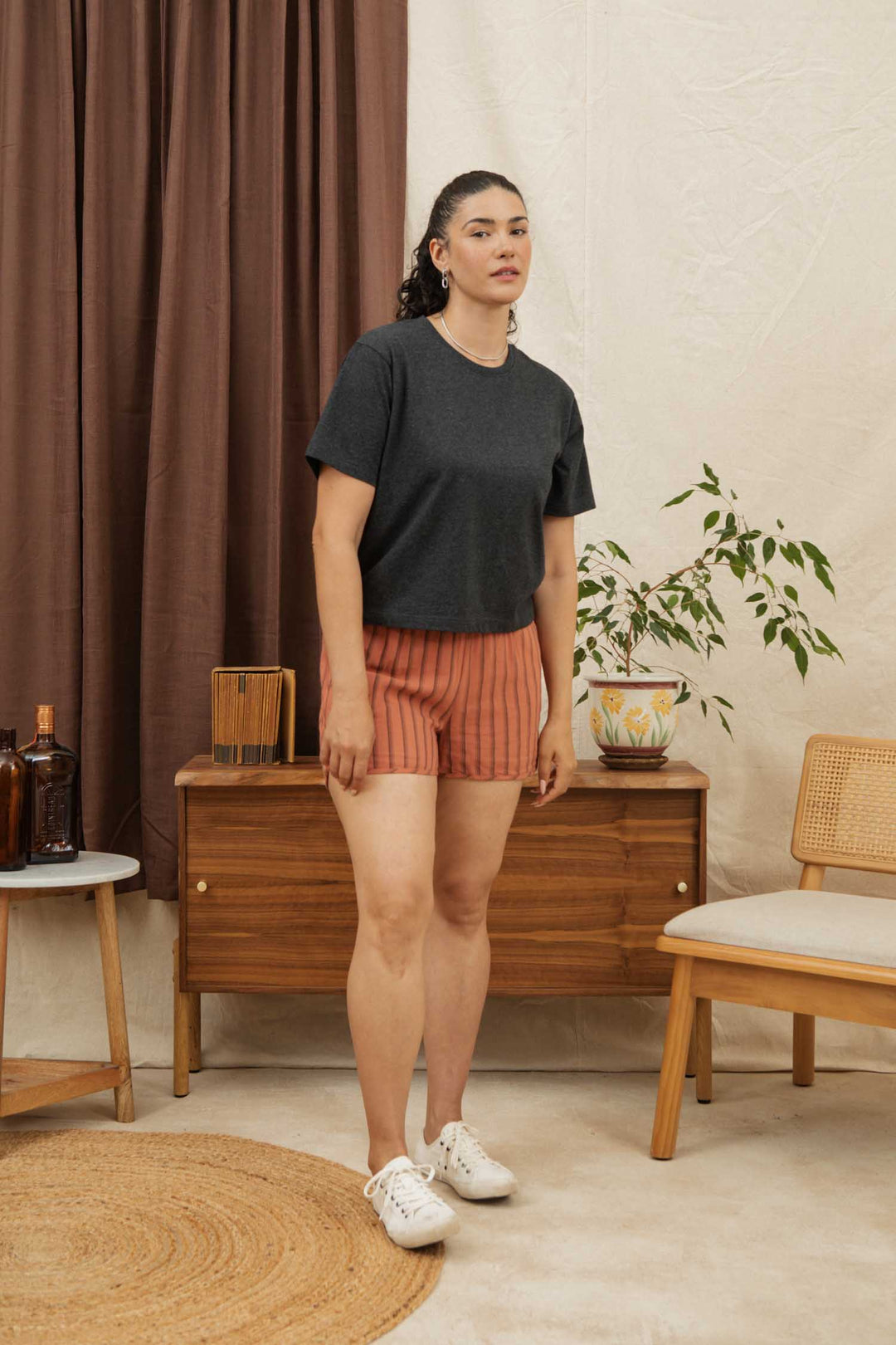 A woman wears the Bridge & Burn Recycled Boxy Tee in Charcoal Heather and orange striped shorts, standing indoors by mid-century furniture, a potted plant, woven mat, and brown curtains, looking at the camera.