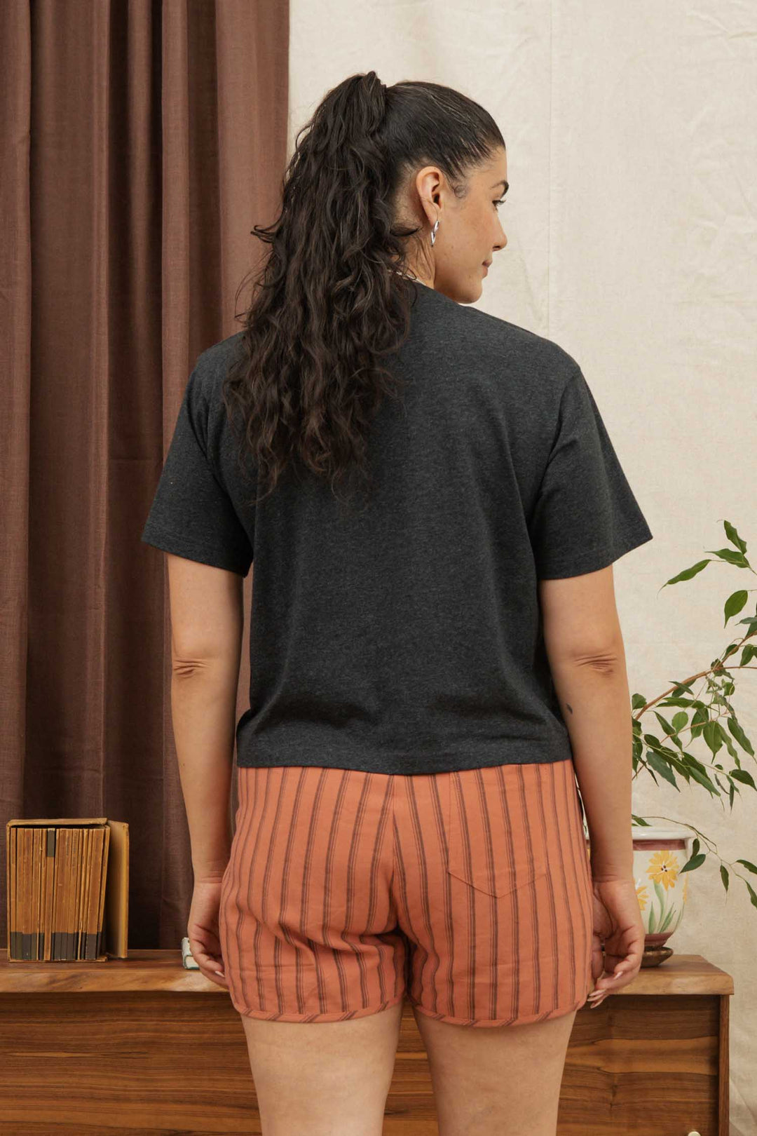 A woman with long, curly hair in a ponytail stands indoors facing away, wearing the Bridge & Burn Recycled Boxy Tee in Charcoal Heather and orange striped shorts. A wooden table, books, plant, and brown curtains appear in the background.