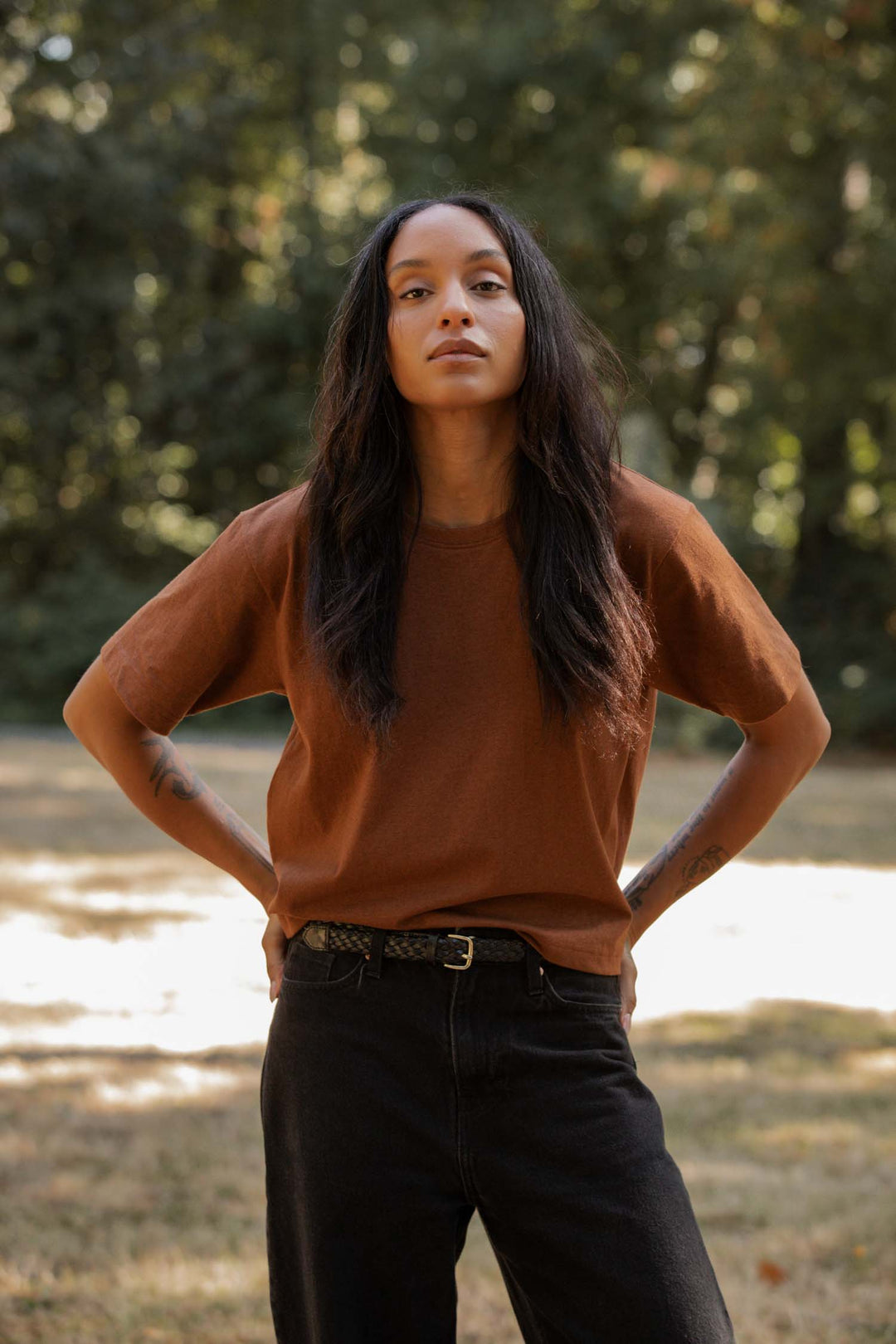 A person with long dark hair stands outdoors, hands on hips, wearing the Bridge & Burn Recycled Boxy Tee in Bronze and black pants. Sunlight filters through trees in the background.