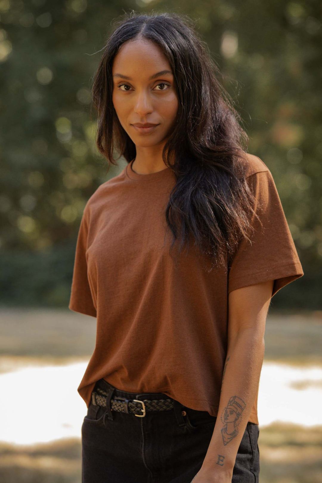 A woman with long dark hair, wearing the Bridge & Burn Recycled Boxy Tee in Bronze and black jeans, stands outdoors amid blurred greenery, looking at the camera. She has a tattoo on her left forearm.