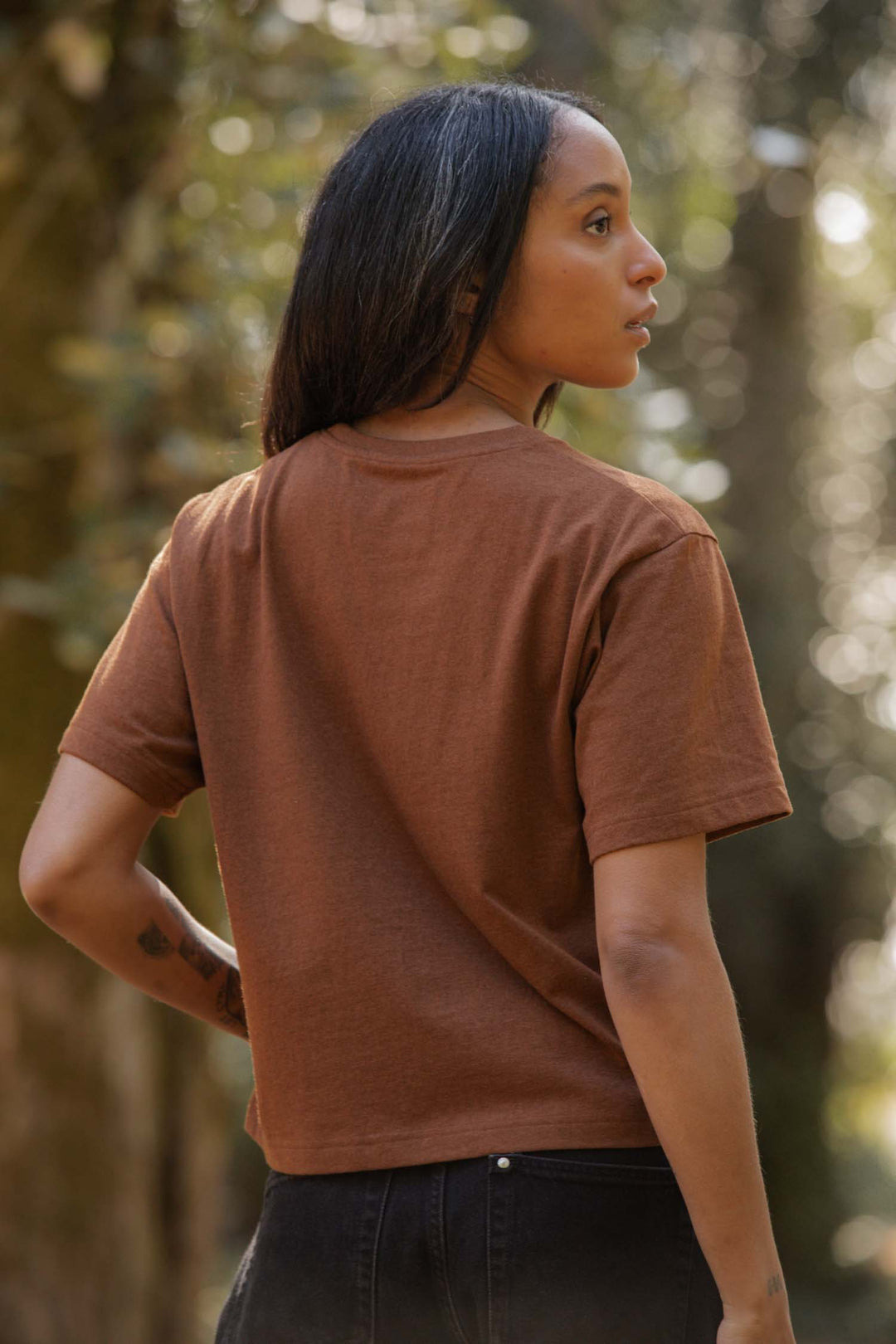 A person with long dark hair, wearing a Bridge & Burn Recycled Boxy Tee in Bronze and dark jeans, stands outdoors with their back turned, looking to the side as sunlight filters through the trees.