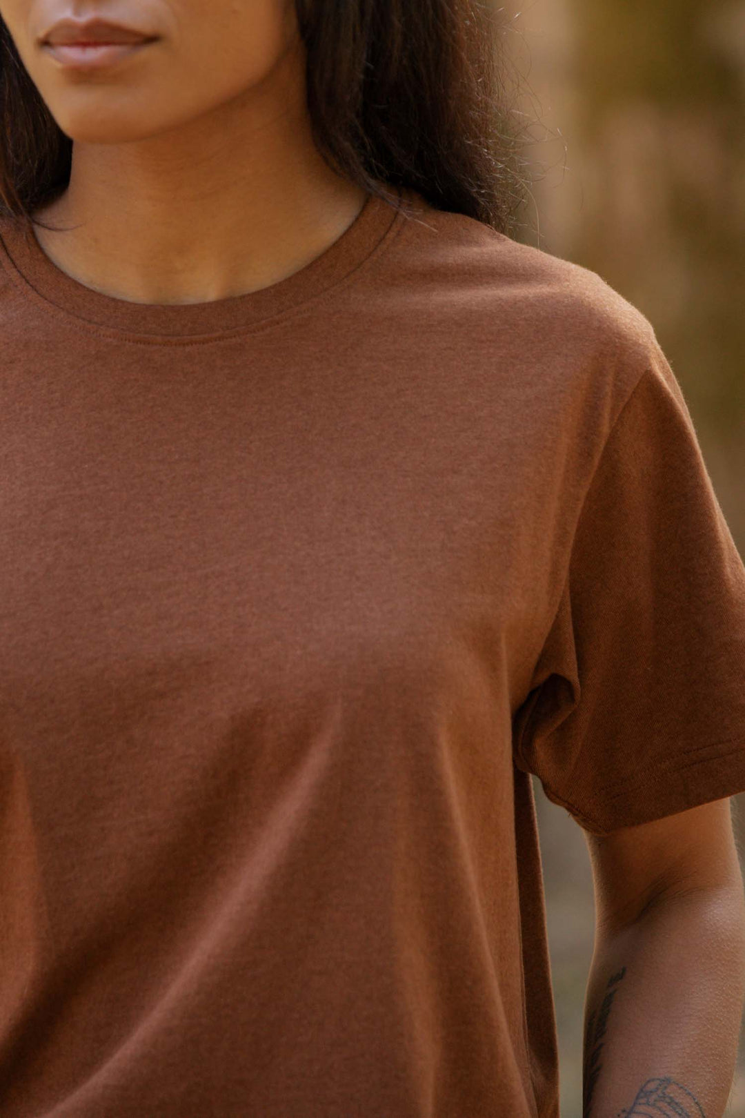 A person with long dark hair is seen outdoors from the shoulders up, wearing the Bridge & Burn Recycled Boxy Tee in Bronze. Their face is partially visible, and a tattoo can be seen on their forearm.