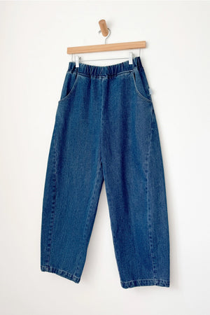 Le Bon Shoppe - Arc Pant / Blue Denim: Relaxed, high-waisted blue denim pants with an elastic waistband and side pockets, shown on a wooden hanger against a white wall.