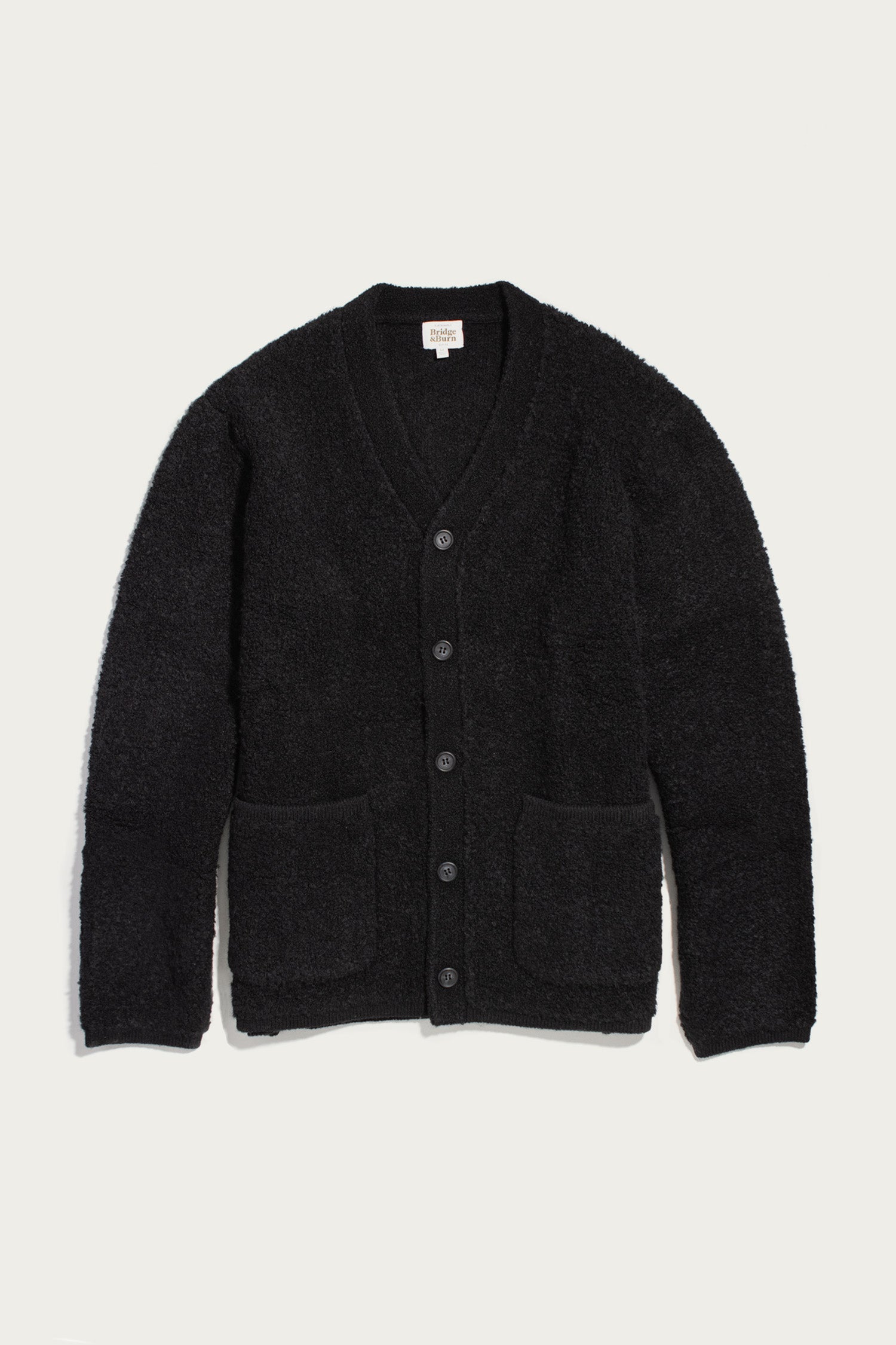The Mateo Cardigan in Black by Bridge & Burn is displayed laid flat on a light background. This V-neck cardigan features patch pockets, a soft textured knit fabric, and a classic button-up design that offers both comfort and style.