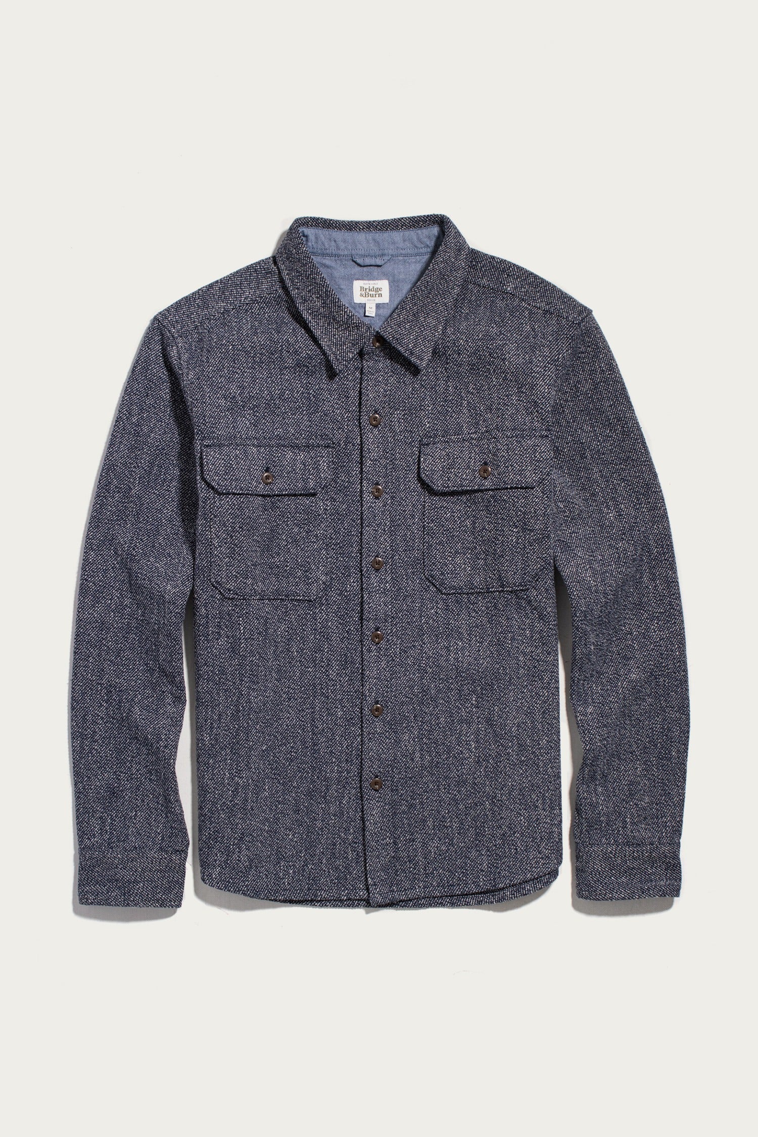 The Bridge & Burn Pointer Overshirt in Ocean Marl—a long-sleeve, button-up blue tweed twill overshirt with two chest pockets and brown buttons—is laid flat on a white background.