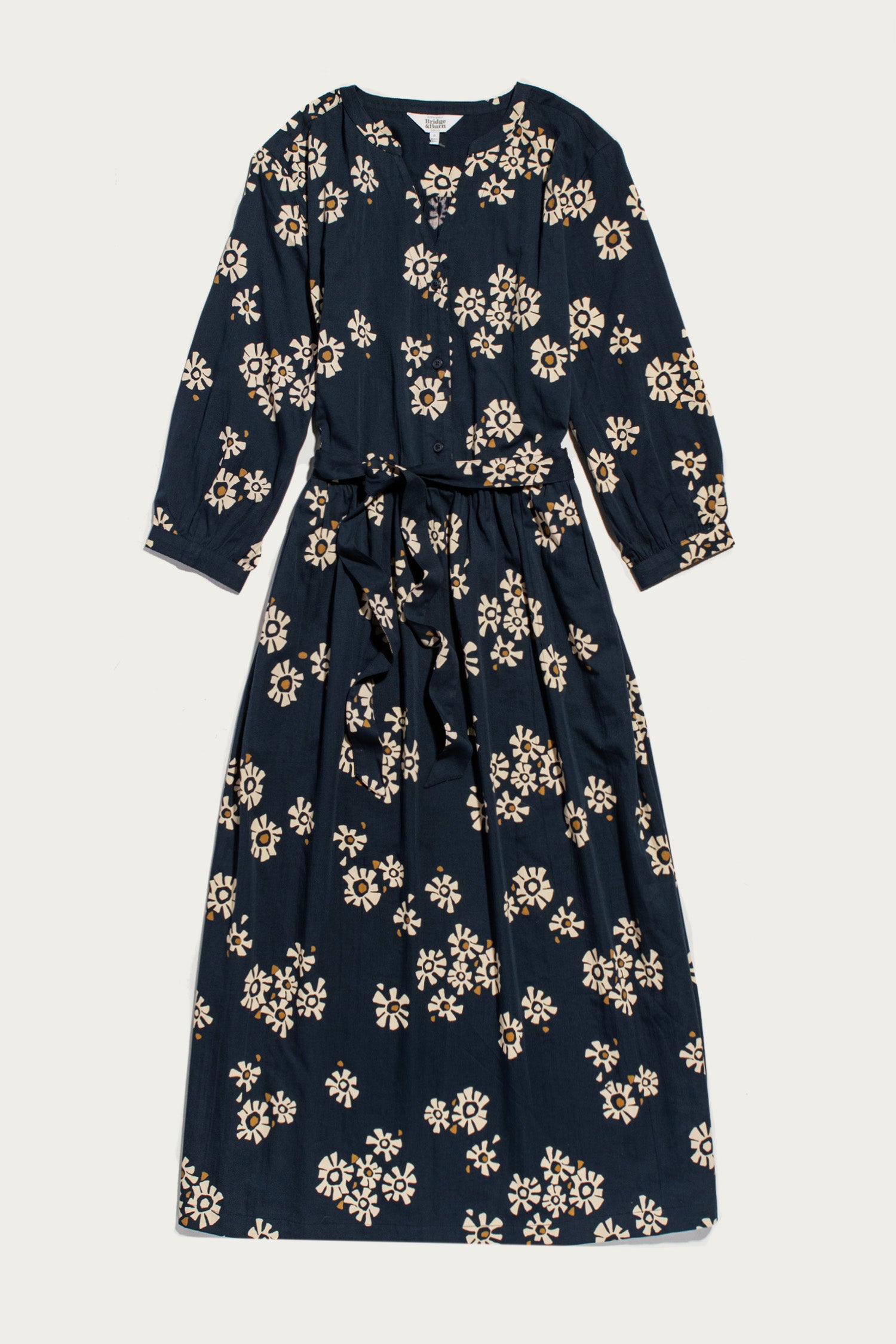The Bridge & Burn Fiona Dress / Night Garden is a long-sleeved midi dress featuring a white and beige floral pattern, gathered sleeves, round neckline, and waist tie, shown laid flat on a light background.