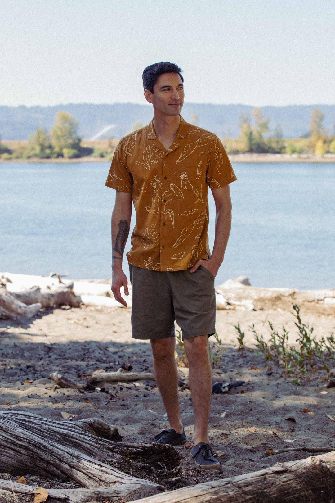 A man stands on a sandy riverbank with trees behind him, wearing the Bridge & Burn Bodie Shirt in Shoreflight, olive shorts, and dark shoes. One hand is in his pocket as he gazes off to the side.