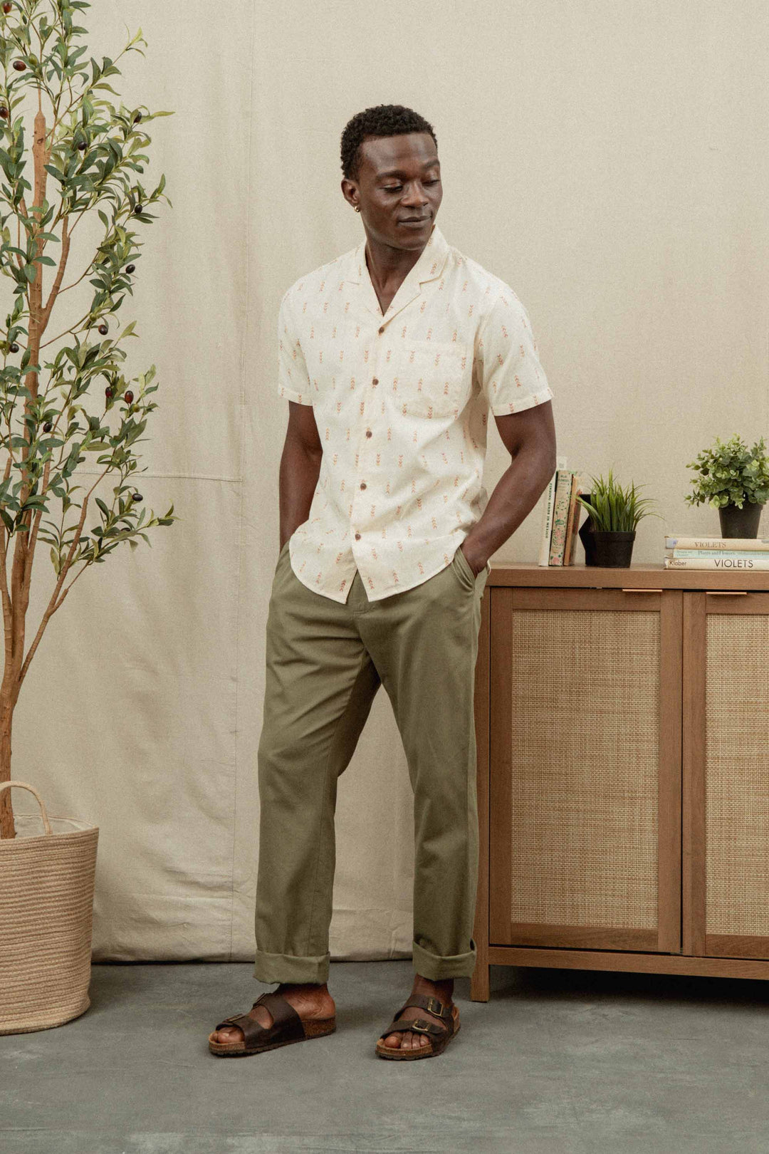 A man stands indoors by a wooden sideboard and potted plant, wearing Bridge & Burn’s Collins Slim Shirt in Dune Chevron, olive green pants, and brown sandals. He has his hands in his pockets and looks slightly down and to the side.