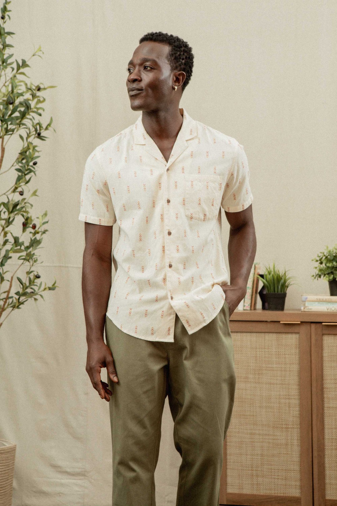 A man stands indoors, smiling with one hand in his pocket, wearing the Bridge & Burn Collins Slim Shirt in Dune Chevron and olive green pants. Plants and a cabinet are visible in the background.
