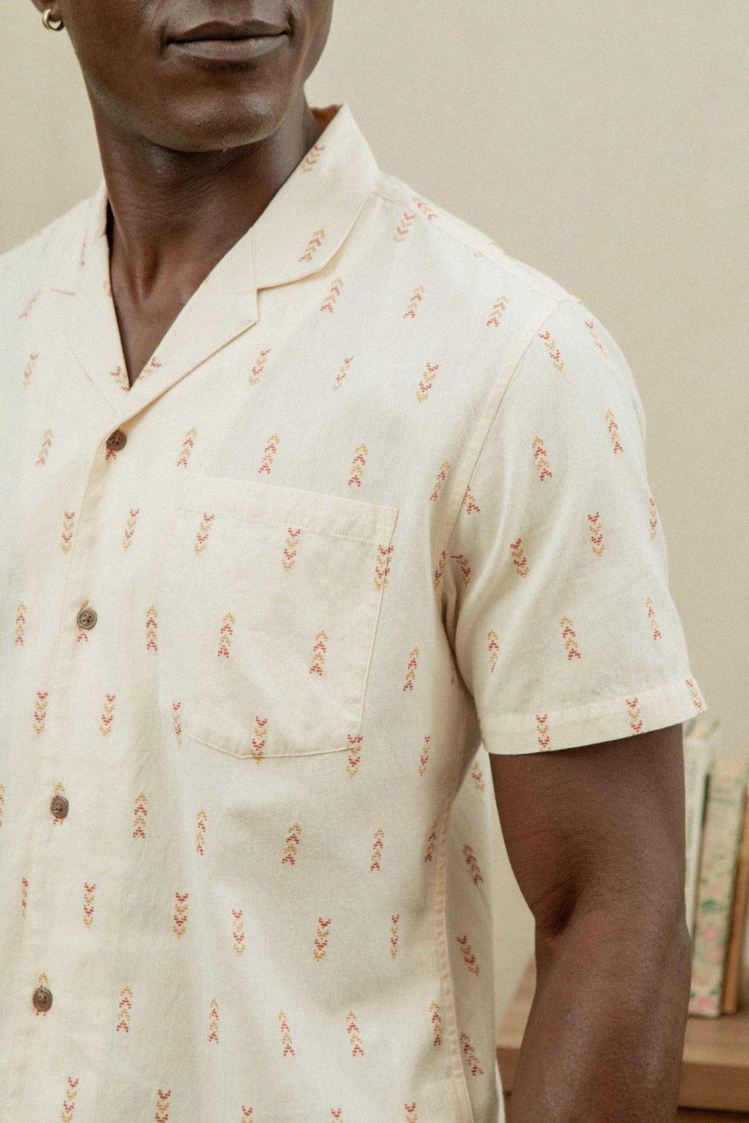 A man wears the Bridge & Burn Collins Slim Shirt in Dune Chevron—a slim fit, cream camp collar shirt with a red pattern, made from lightweight cotton—standing indoors with books blurred in the background.