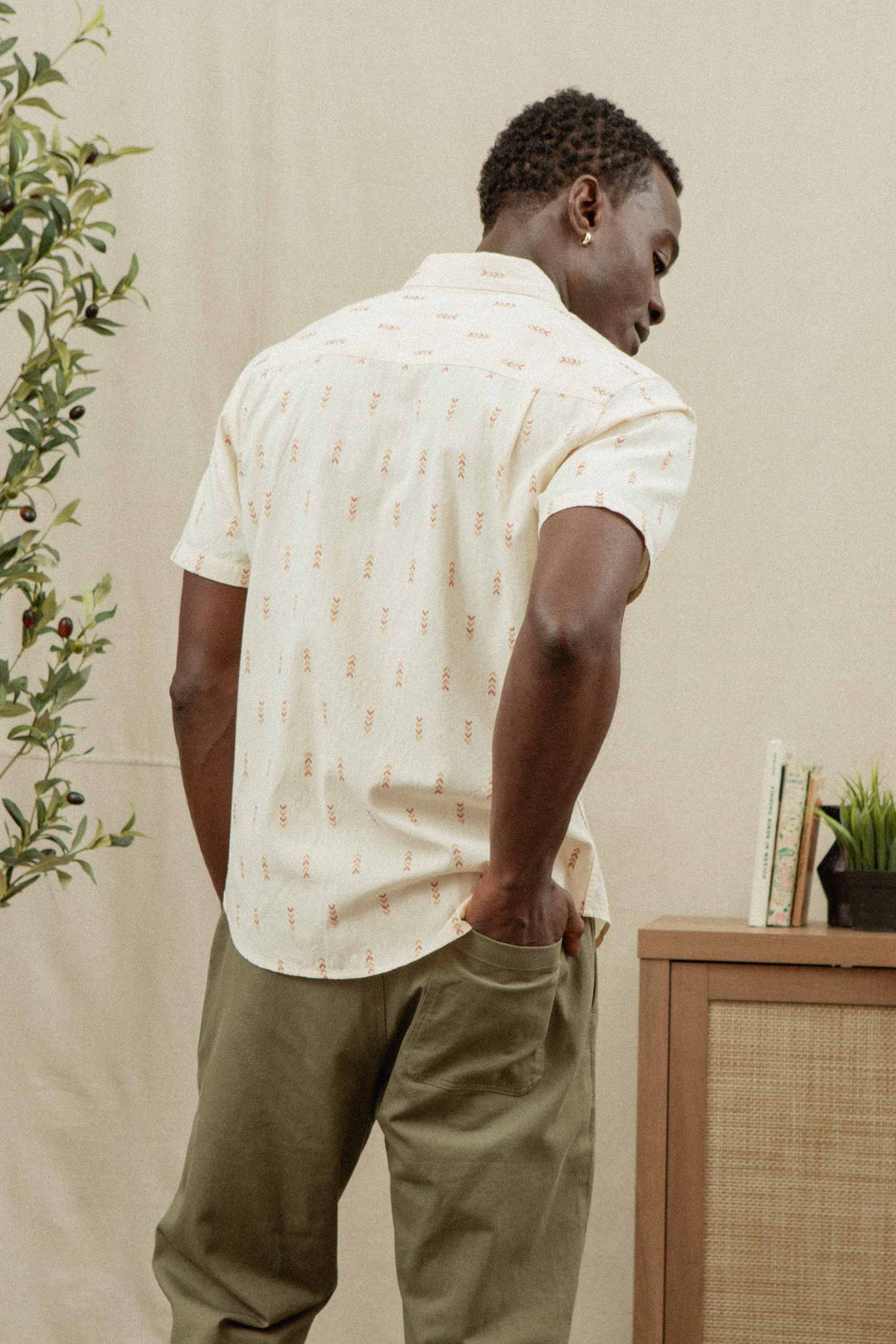 A man with his back to the camera wears a Bridge & Burn Collins Slim Shirt in Dune Chevron and slim fit olive green pants, one hand in his pocket. Plants and a bookshelf are visible in the background.