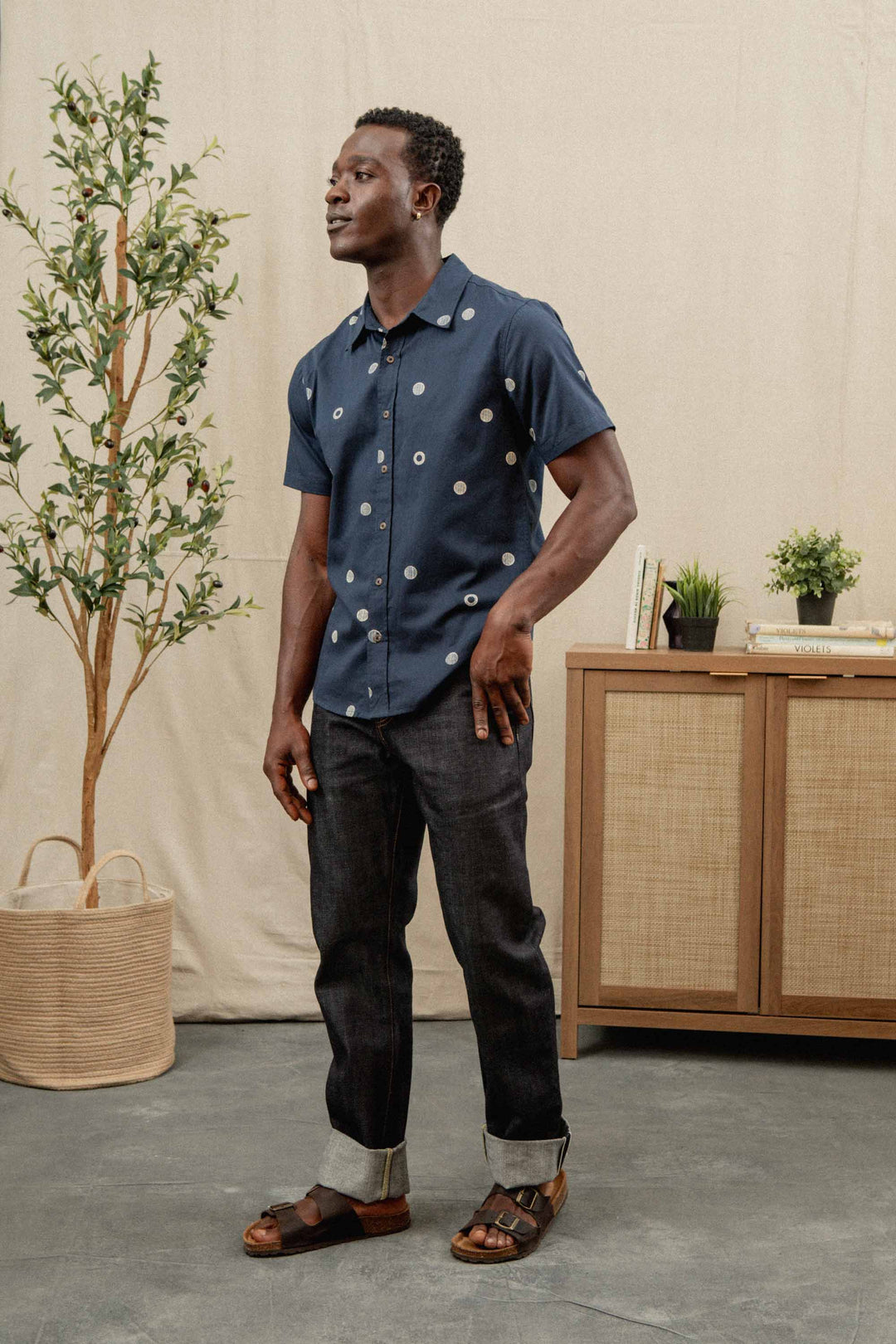 A man stands indoors on a gray floor wearing the Bridge & Burn Emerson Slim Shirt in Lunar Polka, dark cuffed jeans, and brown sandals. Behind him are a potted plant and a wooden cabinet with books and plants.