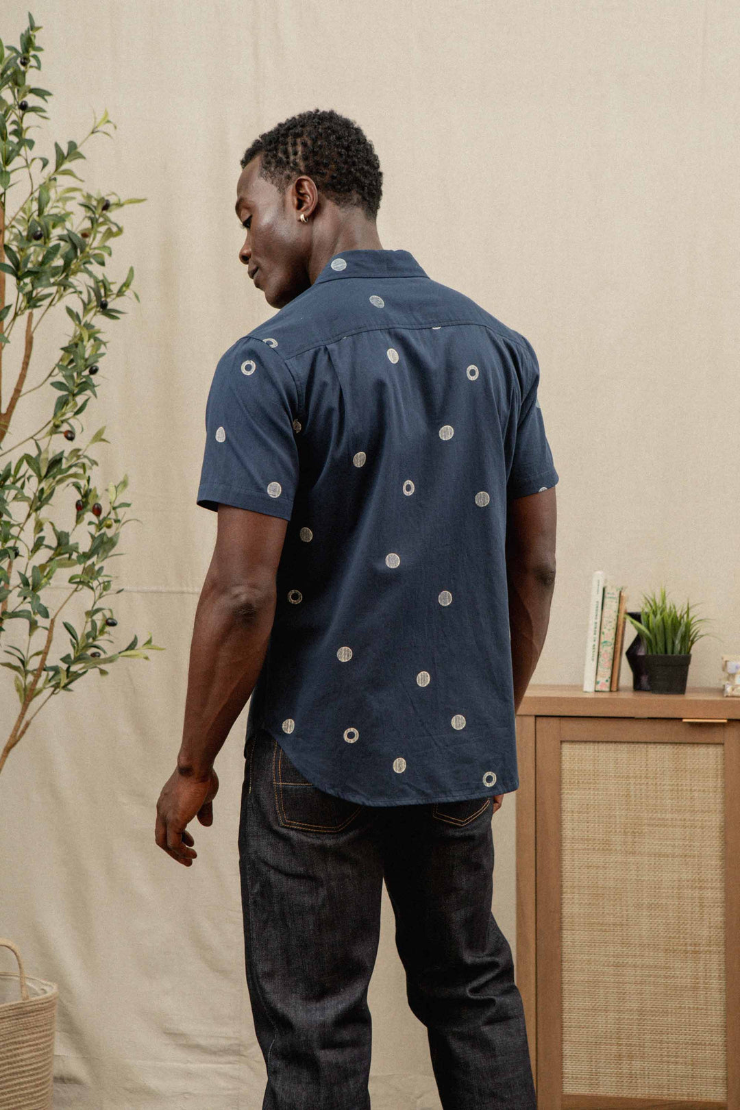 A man wearing the Bridge & Burn Emerson Slim Shirt in Lunar Polka and dark jeans stands with his back to the camera in a cozy room with a plant and wooden cabinet.