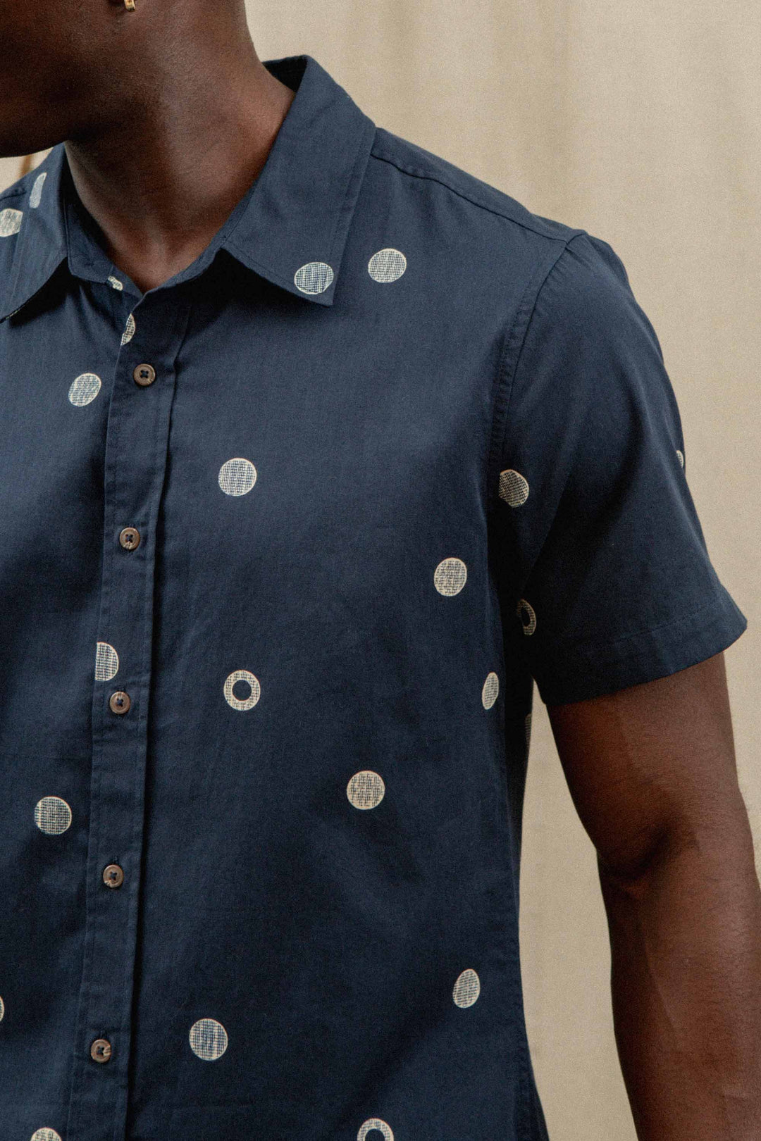 A person wears the Bridge & Burn Emerson Slim Shirt in Lunar Polka—a navy, short-sleeve, slim fit button-up with light circular dots—against a neutral background, showing only their upper body.
