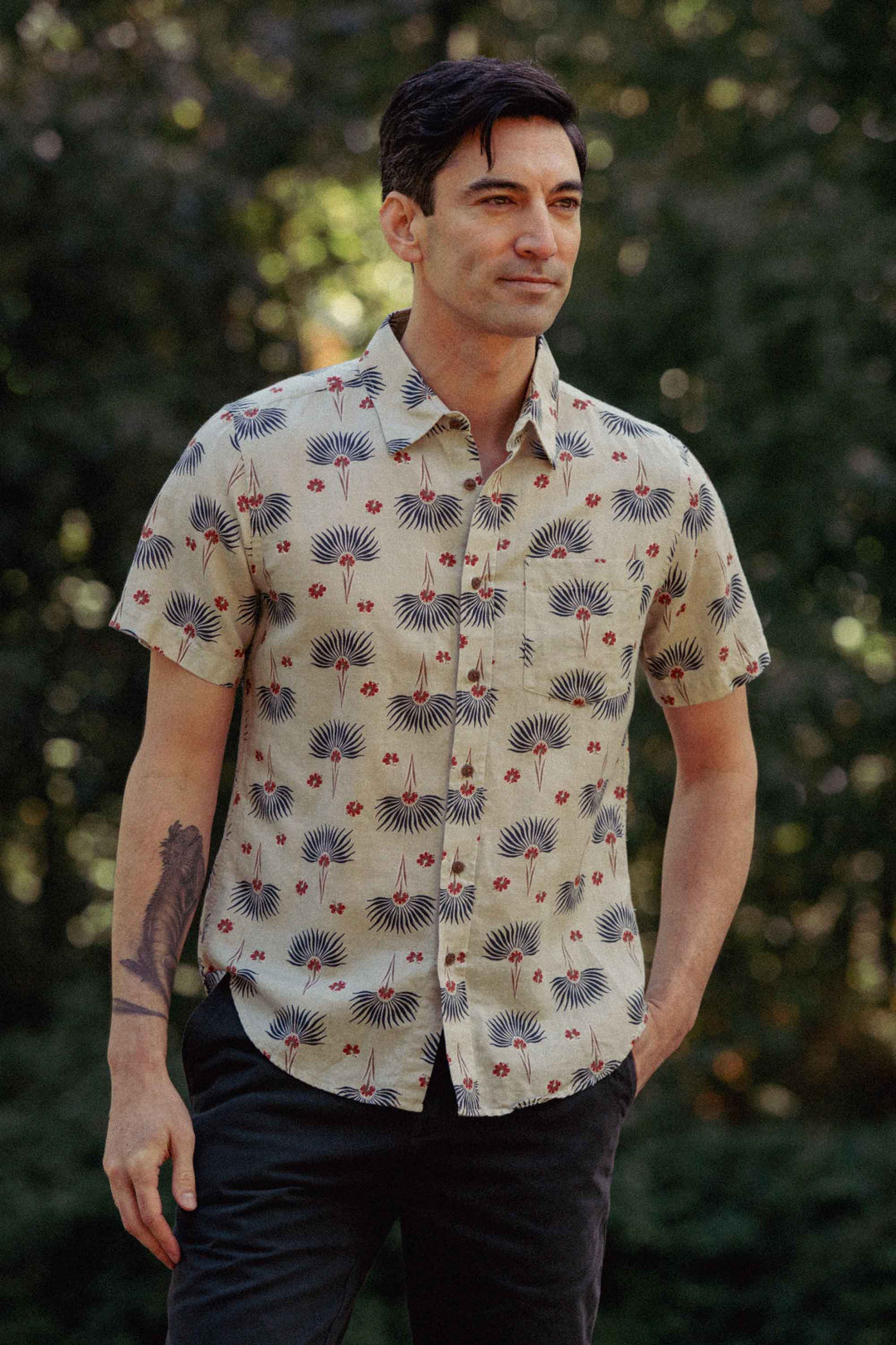 A man with dark hair wears the Bridge & Burn Harbor Slim Shirt in Aloha Floral, standing outdoors with one hand in his pocket against a blurred green background.
