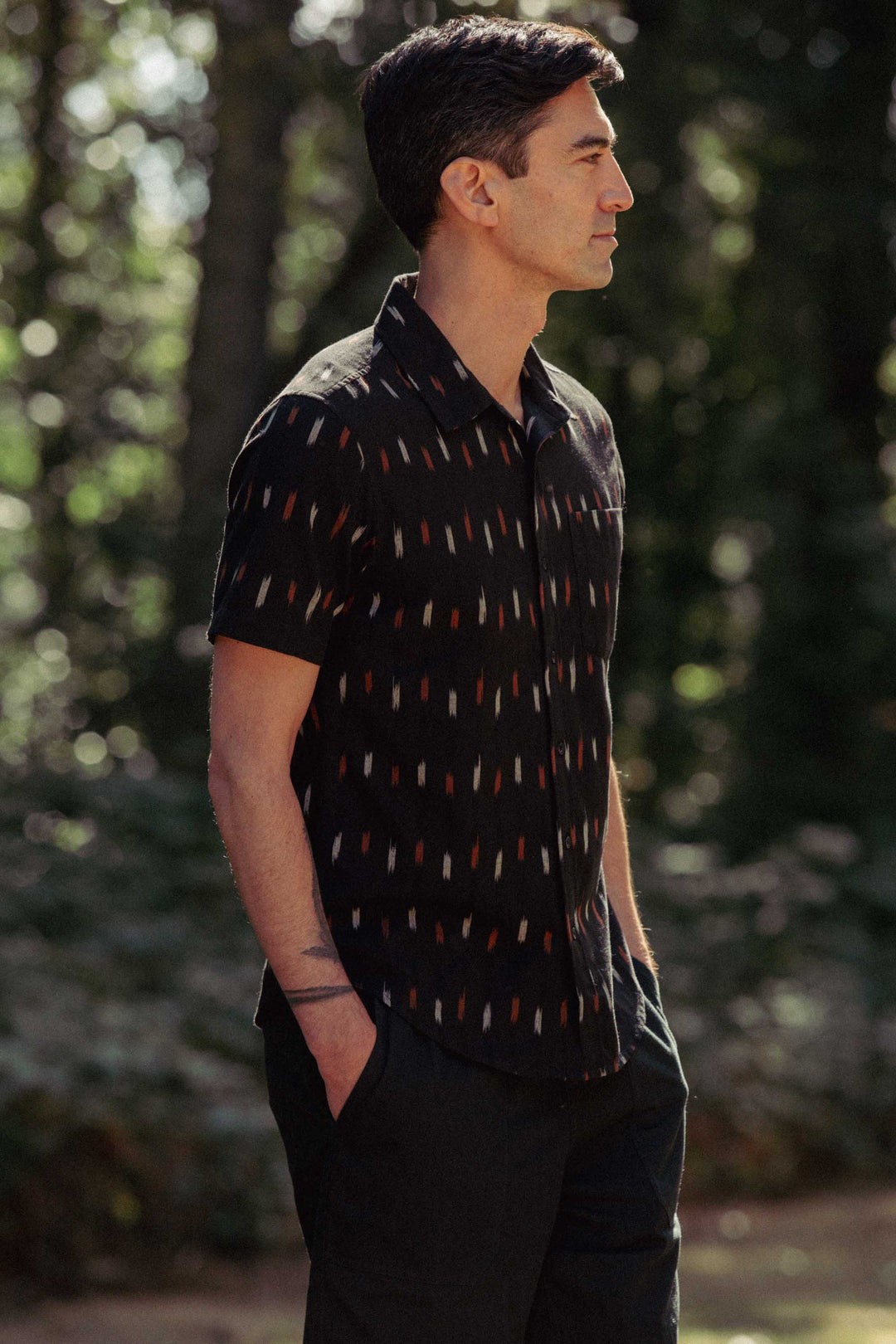 A man with short dark hair stands outdoors in profile, hands in pockets, wearing the Bridge & Burn Harbor Slim Shirt in Dotted Horizon and black pants. Trees and greenery create a blurred background.