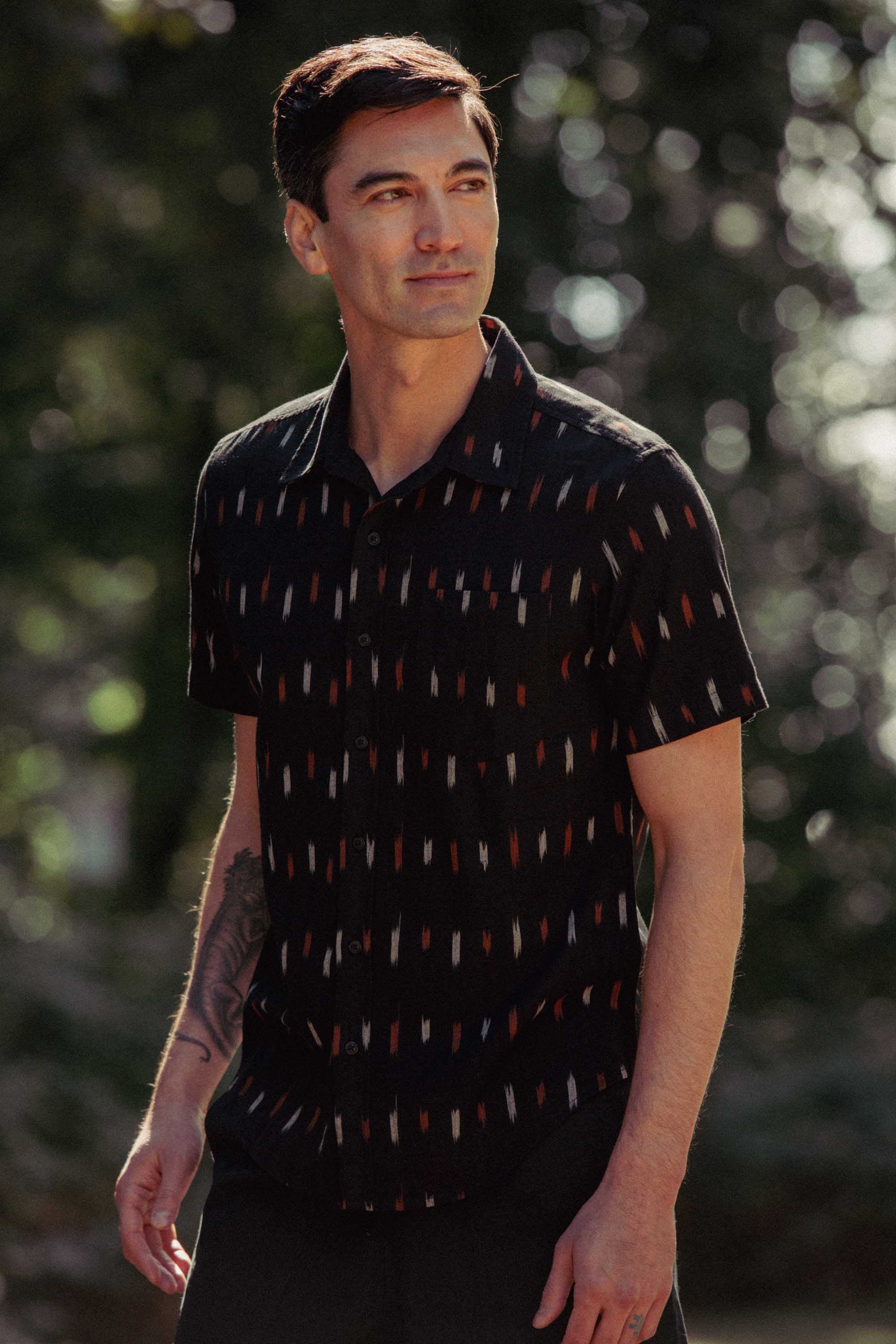 A man with short dark hair stands outdoors in the Bridge & Burn Harbor Slim Shirt in Dotted Horizon, a black summerweight cotton shirt with a geometric pattern, showing off his tattooed forearm against blurred greenery.