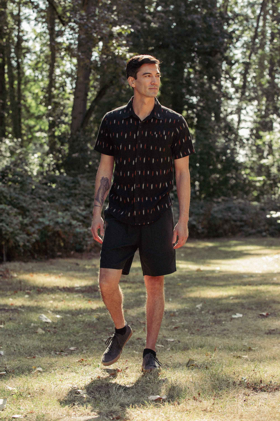 A man walks on grass in sunlight wearing the Bridge & Burn Harbor Slim Shirt in Dotted Horizon, paired with black shorts and sneakers. Tall trees stand in the background.
