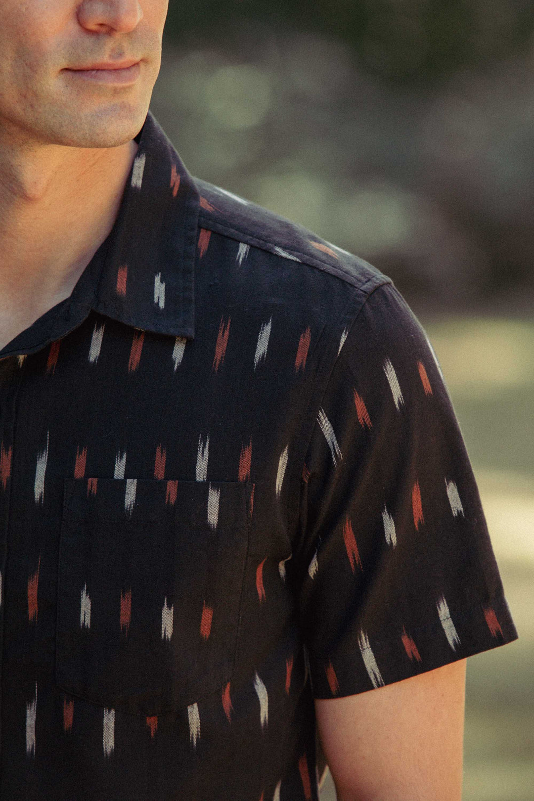 A close-up of a man wearing the Bridge & Burn Harbor Slim Shirt in Dotted Horizon—a versatile, slim-fit short sleeve shirt with a white and brown geometric pattern; his face is partly visible against a blurred background.