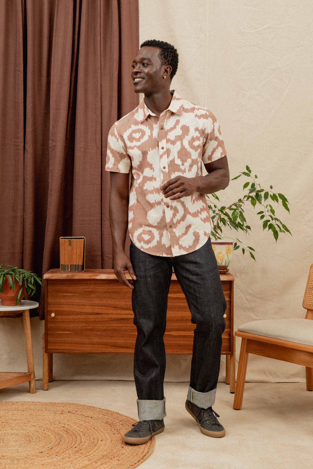 A man stands indoors smiling and looking to the side, wearing the Bridge & Burn Harbor Slim Shirt in Pop Leaf and dark cuffed jeans. He is next to a mid-century wooden console, a potted plant, and a brown curtain.