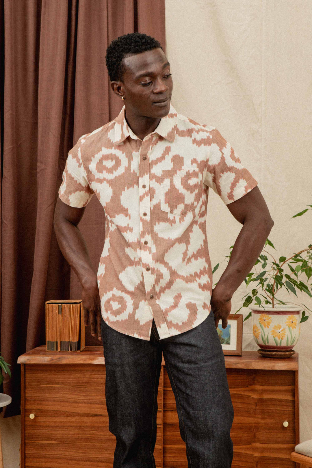 A man wears the Bridge & Burn Harbor Slim Shirt in Pop Leaf, styled with dark jeans, standing indoors before a wooden dresser decorated with plants and decor items.