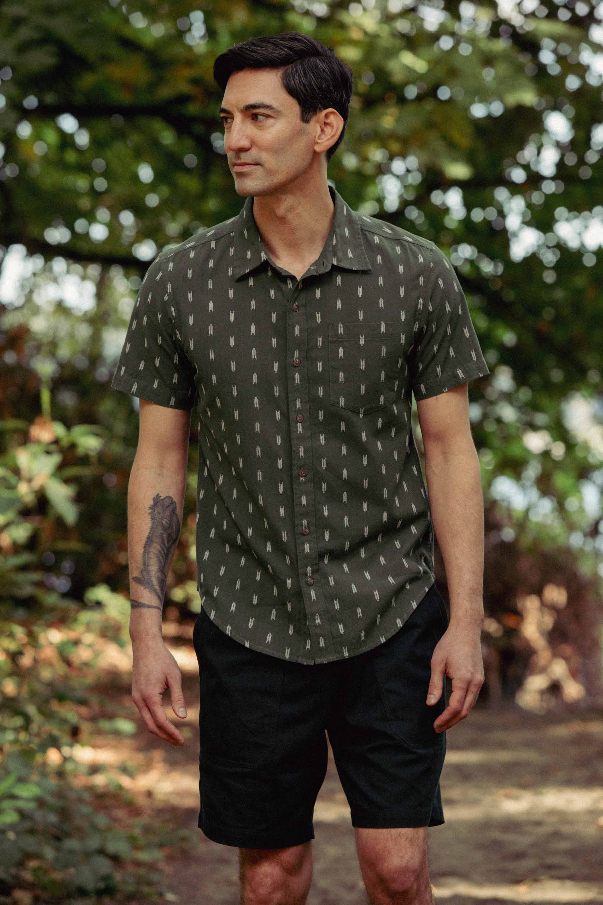 A man with short dark hair and an arm tattoo stands among lush greenery, wearing the Bridge & Burn Harbor Slim Shirt in Thyme Arrows and black shorts. The tailored fit shirt features a subtle pattern, perfect for summer.
